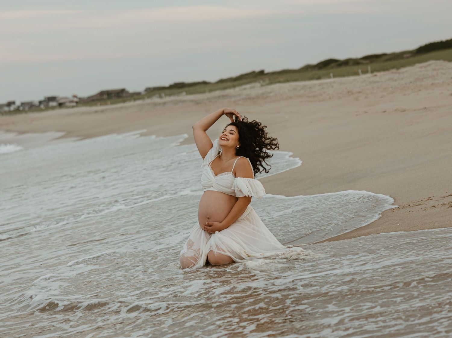 Pregnant woman in white dress kneeling in ocean waves on sandy beach during golden hour maternity photoshoot.