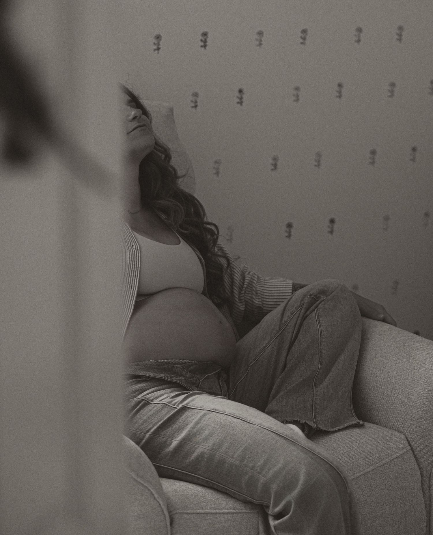 Black and white photo of a pregnant woman resting on a couch, looking up at a wall with small decorations.