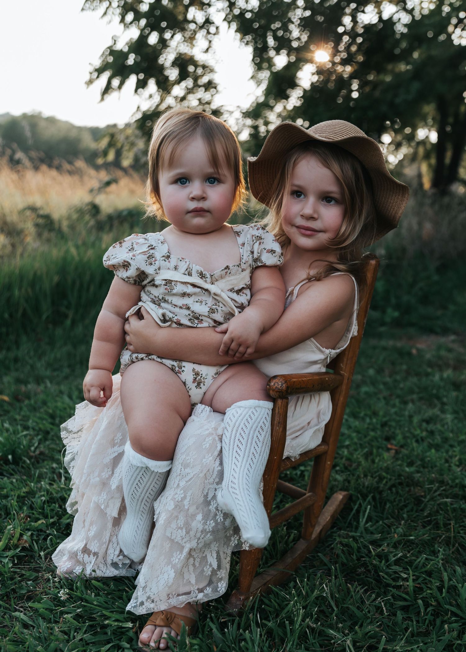 Children sitting together in rustic chair outdoors wearing white lace dresses.