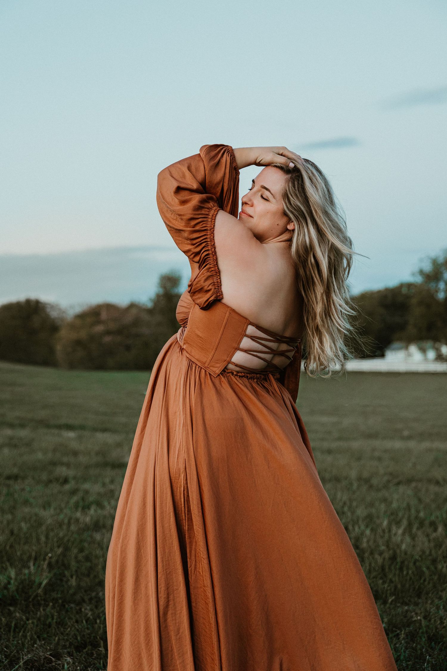 Woman in backless rust orange gown poses gracefully in green field at dusk with hair flowing.