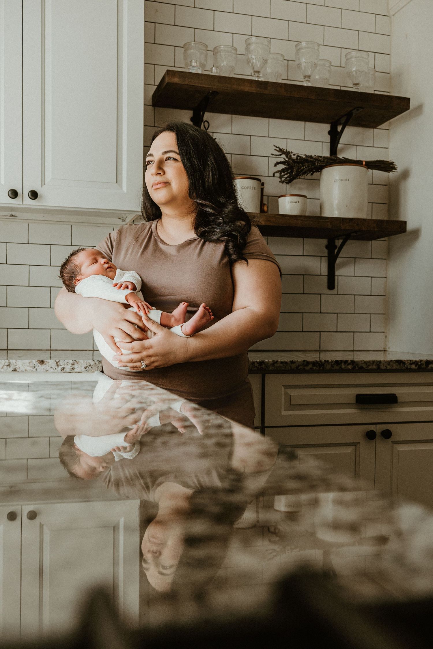 A mother tenderly holds her newborn baby in a modern white kitchen with subway tile backsplash and wooden shelves.