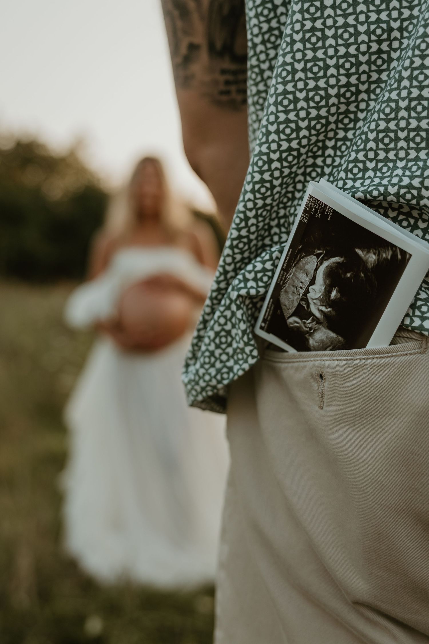 Man with tattoo holding ultrasound photo in pocket, pregnant woman in white dress visible in background outdoors.