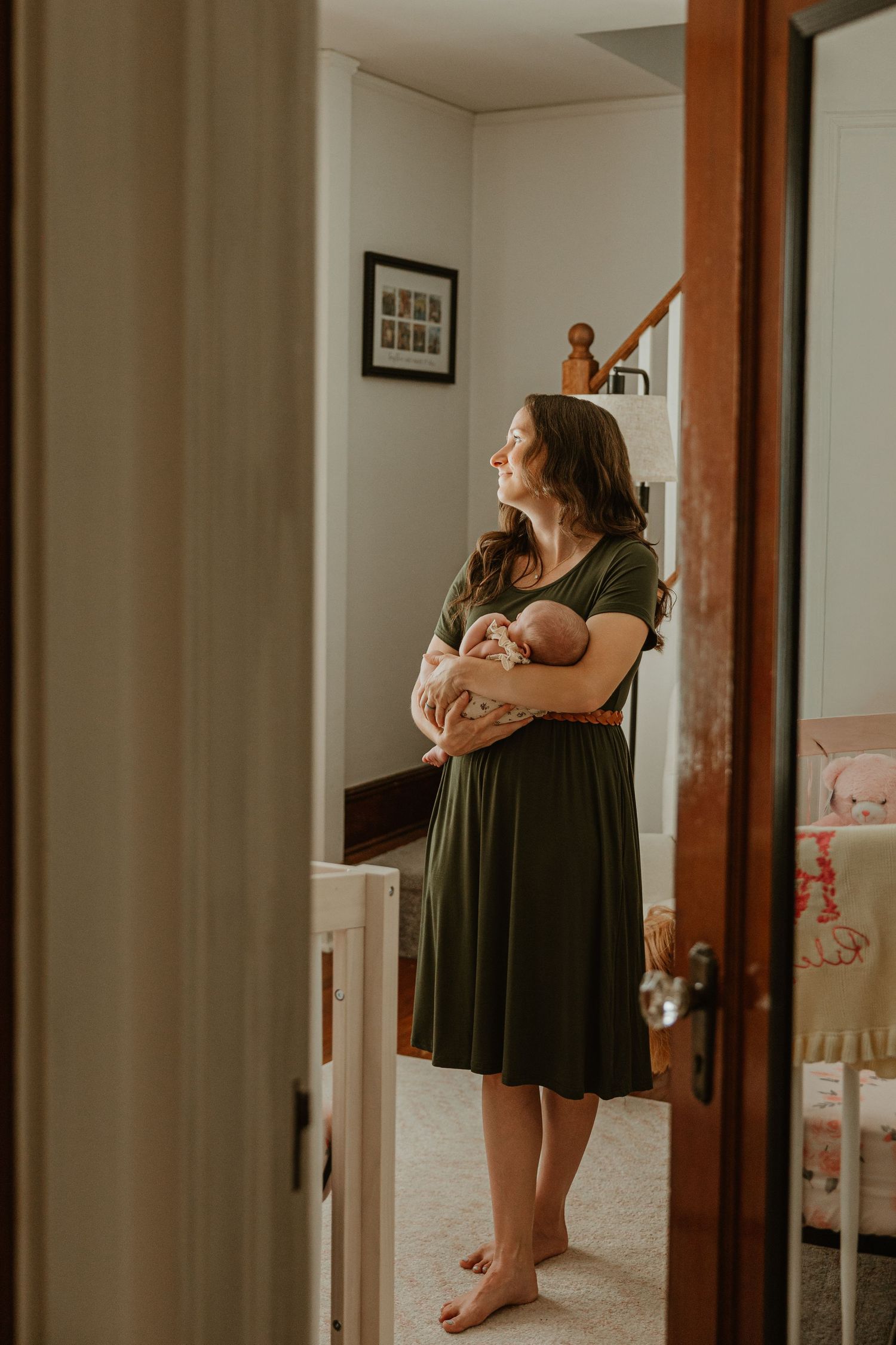 A mother in a green dress holds her newborn baby while standing barefoot in a softly lit nursery room.