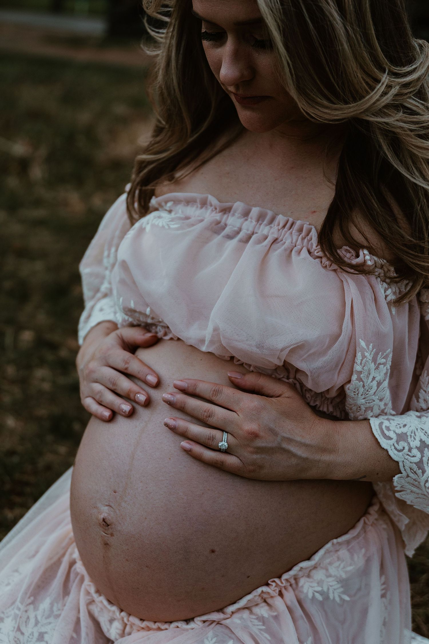 Moody maternity portrait of woman in pink lace dress tenderly holding her baby bump.