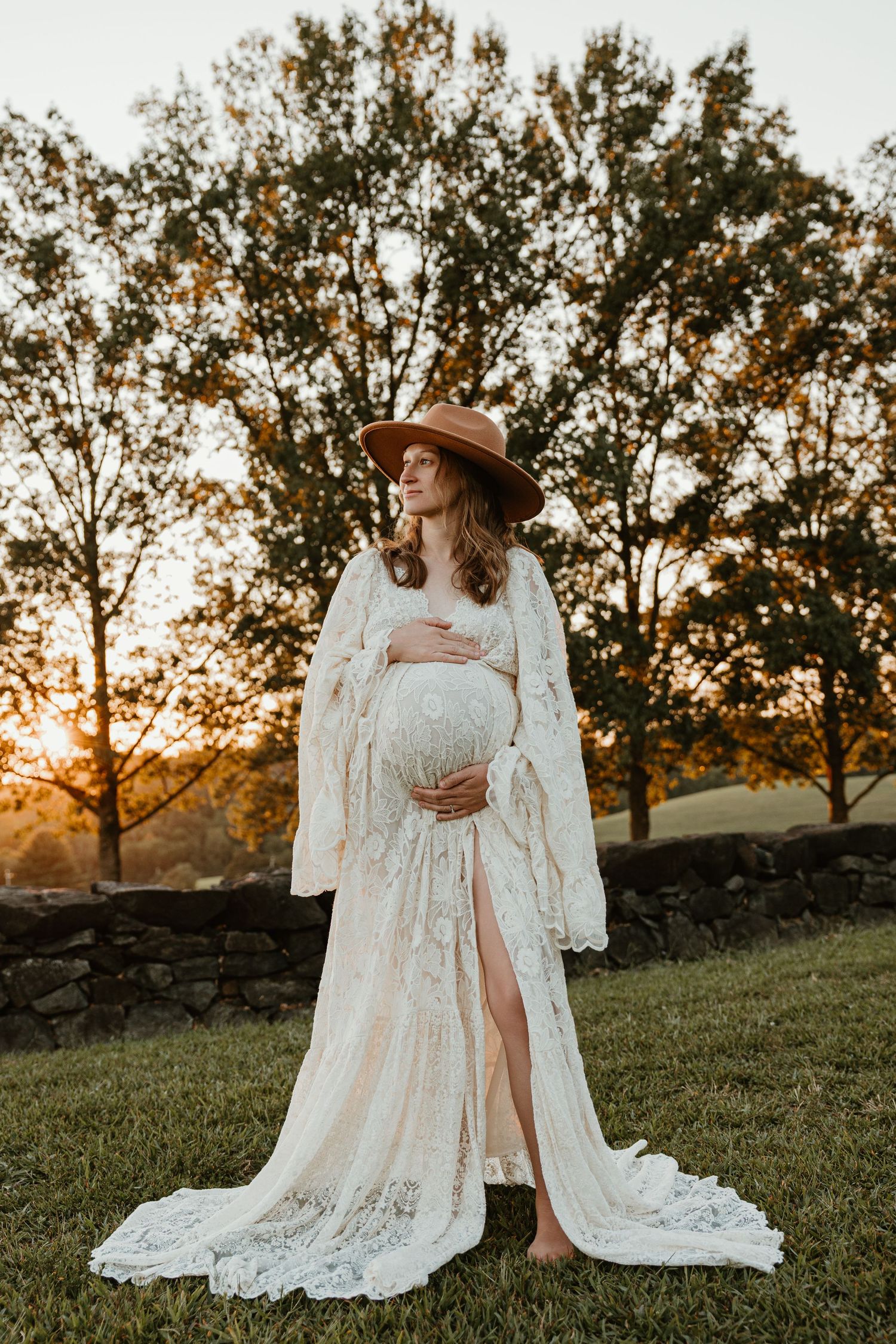 Ethereal maternity photos in a flowing white lace dress against sunset and stone wall backdrop.