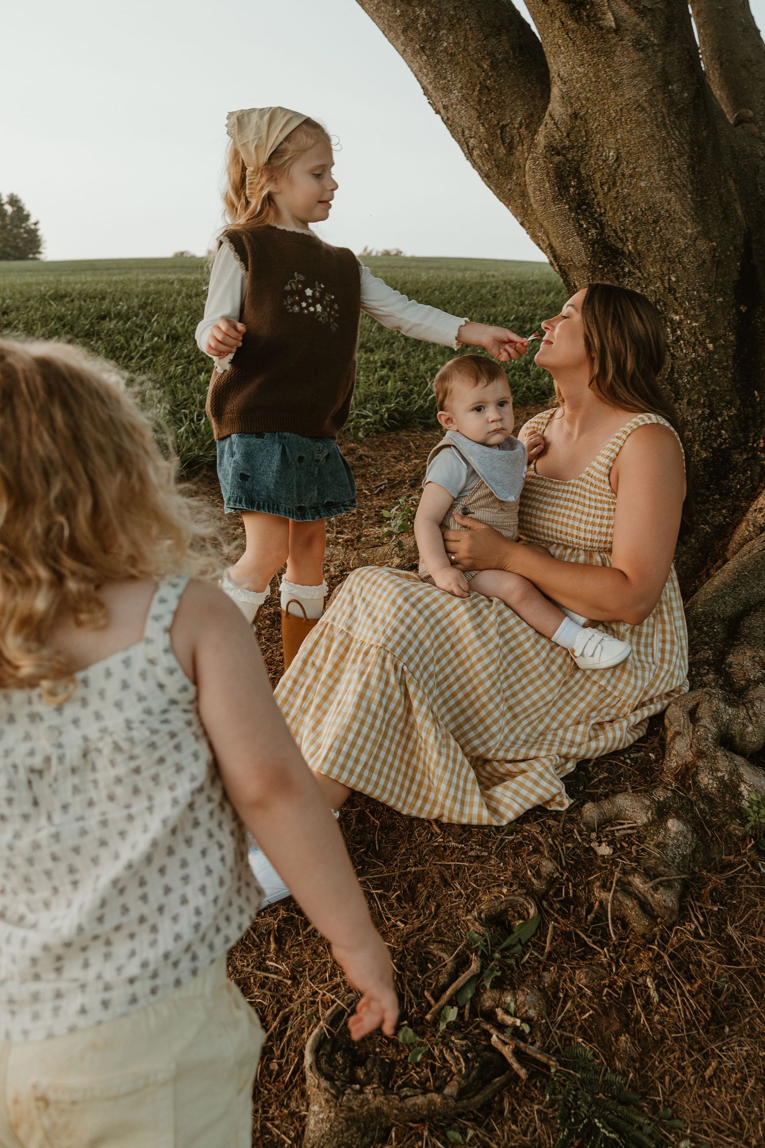 A mother sits under a tree holding a baby while two young girls play around her in a green field.