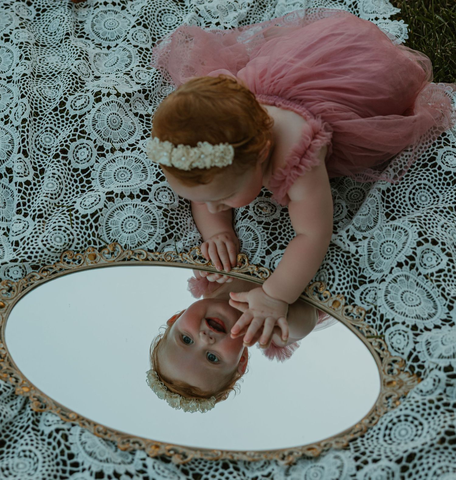 Baby girl in pink tutu dress looks at her reflection in an oval mirror on lace fabric outdoors.