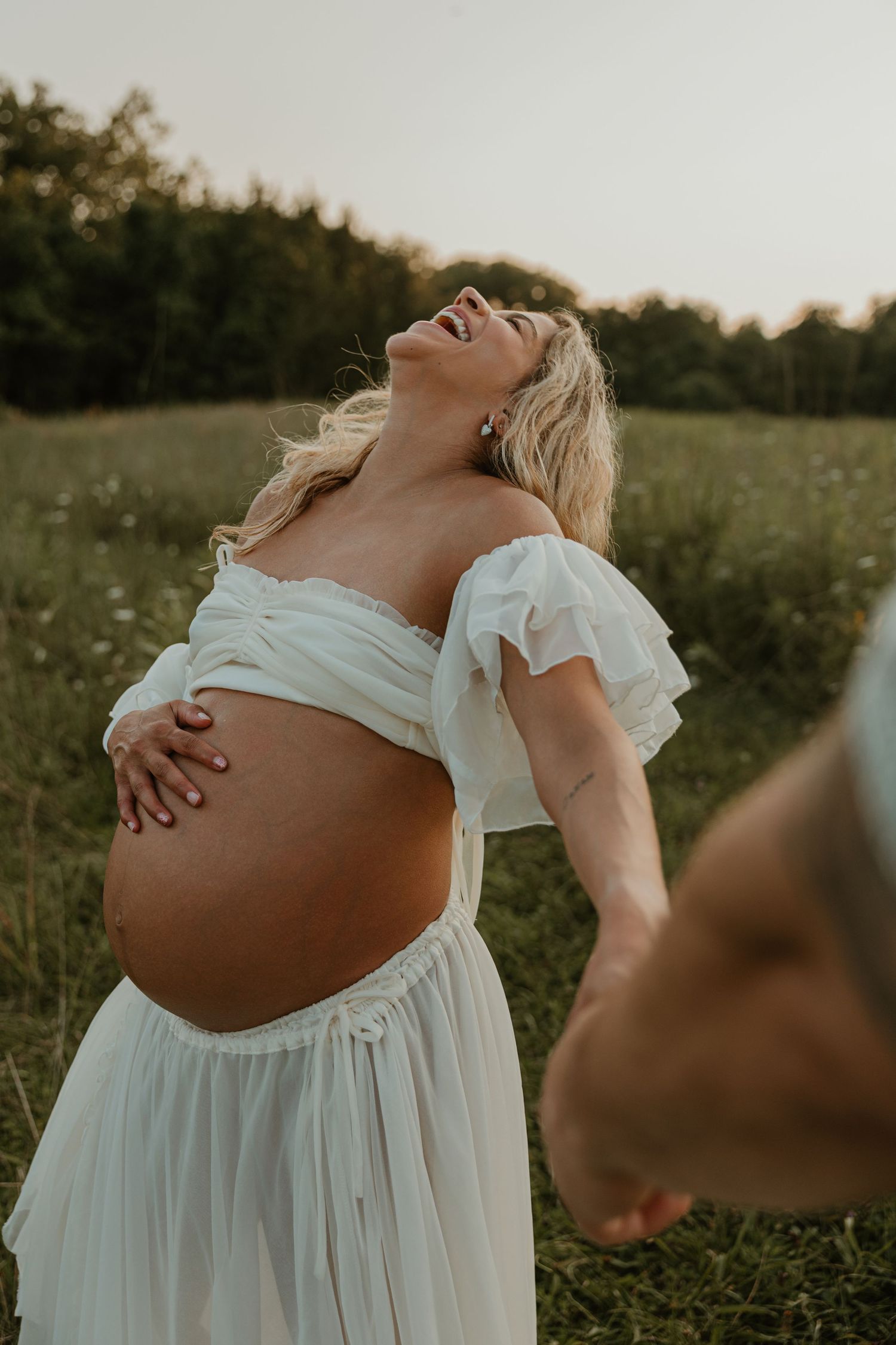 Pregnant woman in white off-shoulder dress laughing joyfully in a green field during golden hour maternity photoshoot.