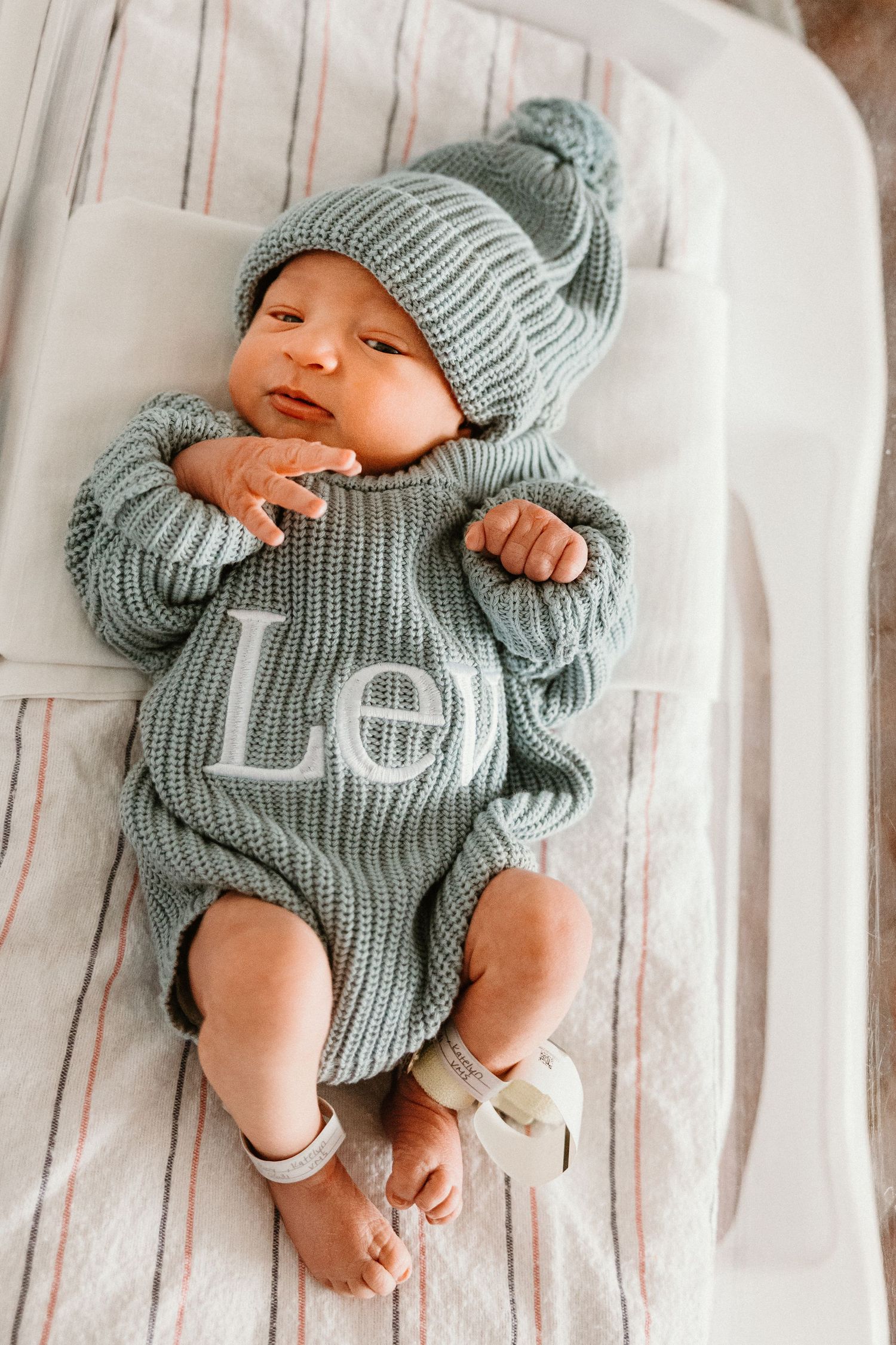 Newborn baby in sage green knit romper and matching pom-pom hat with monogram letters, lying on white bedding.