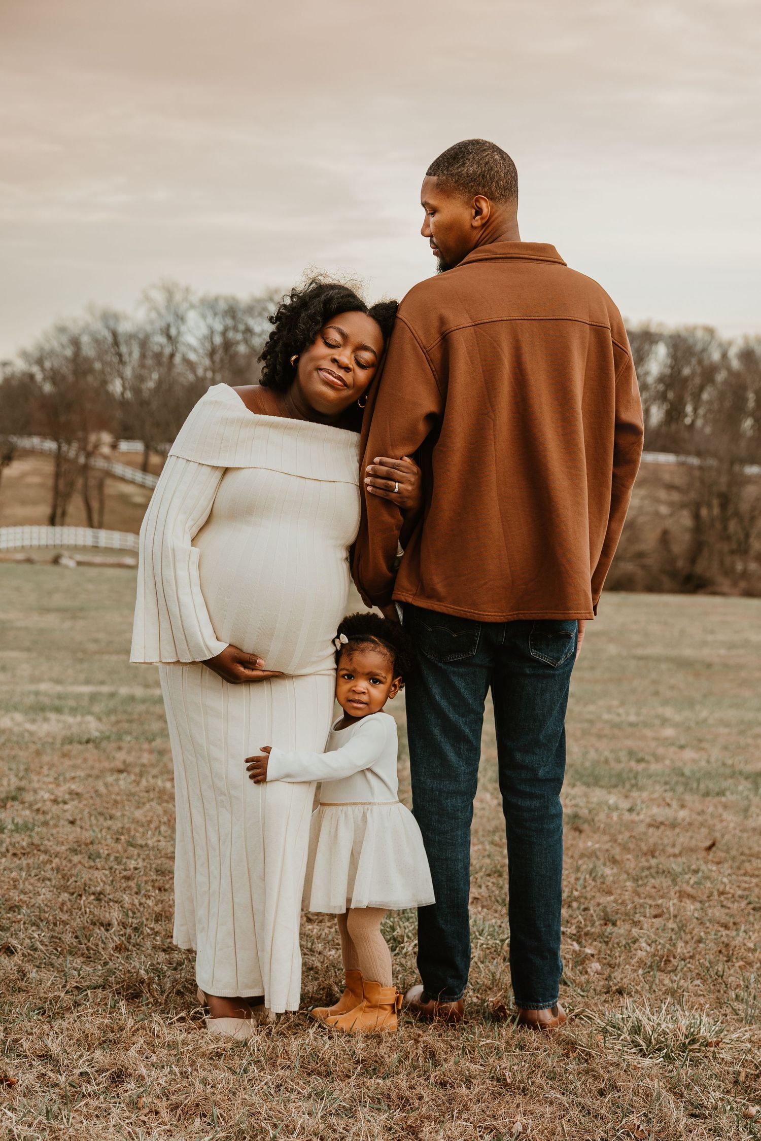 Pregnant Black woman in white dress leans on partner outdoors as toddler daughter hugs her, family maternity photo session.