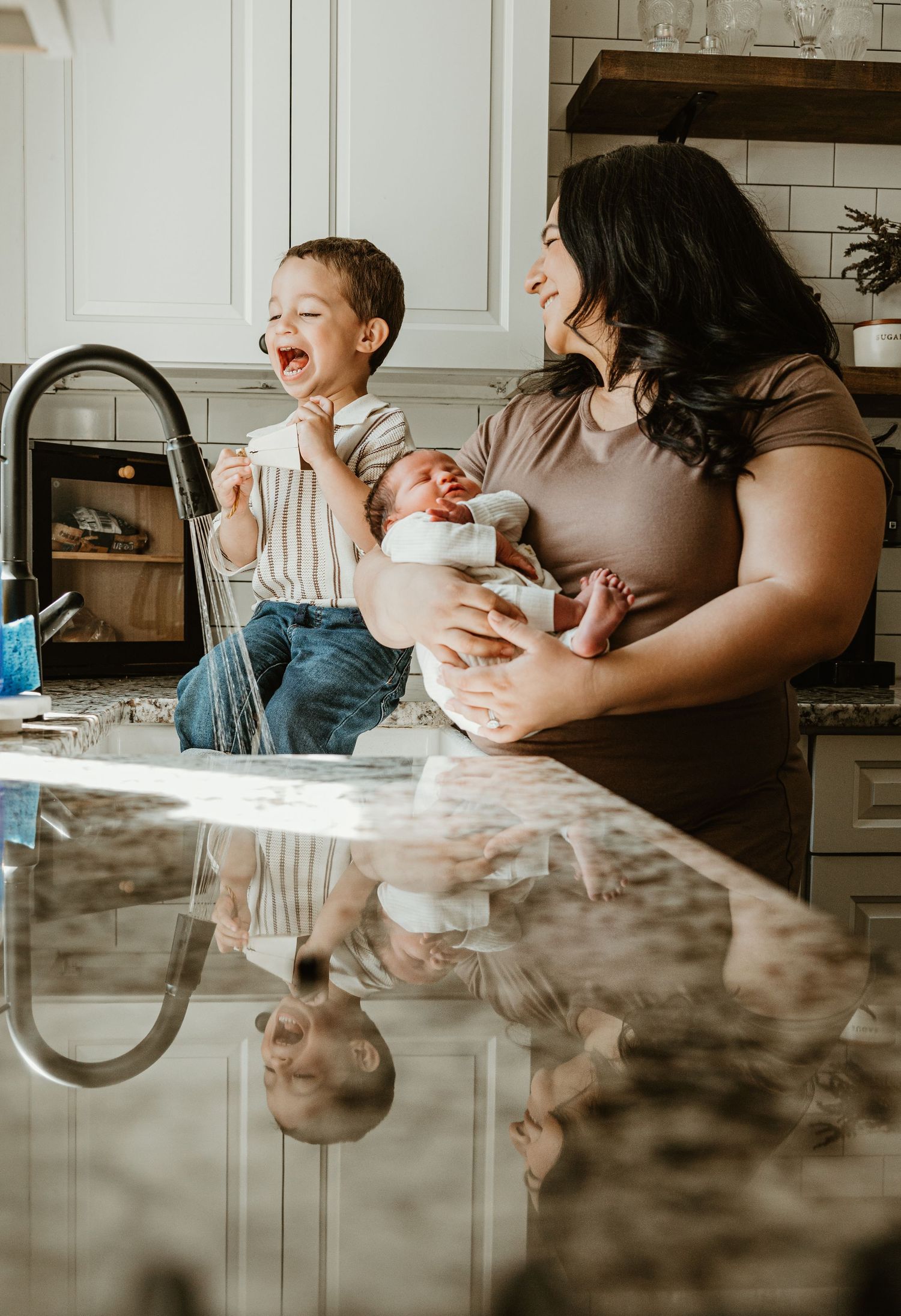 Happy mother holding newborn baby while laughing toddler son sits on kitchen counter beside her.