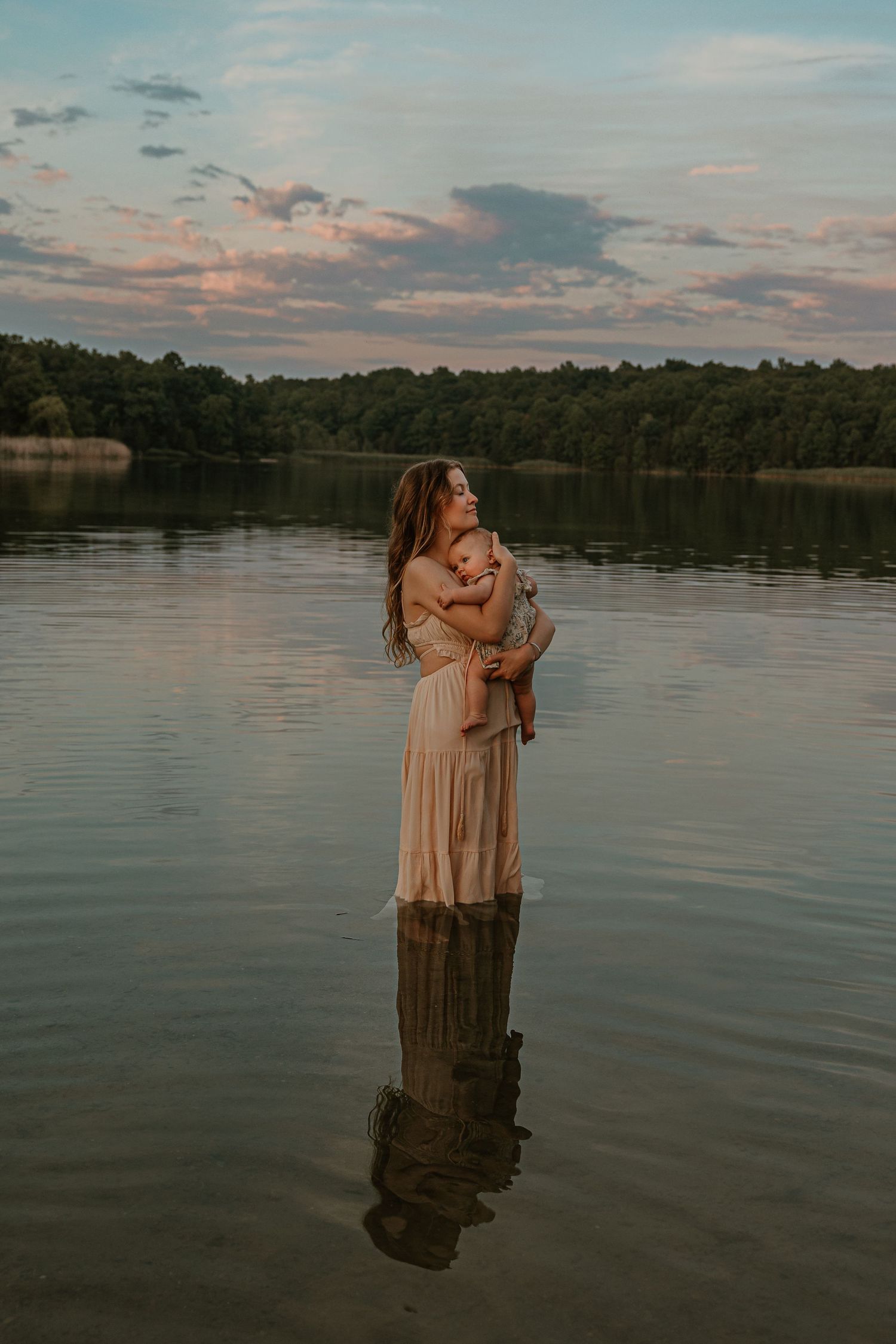 Woman in beige dress holding baby while standing in calm lake at sunset with lush trees in background.