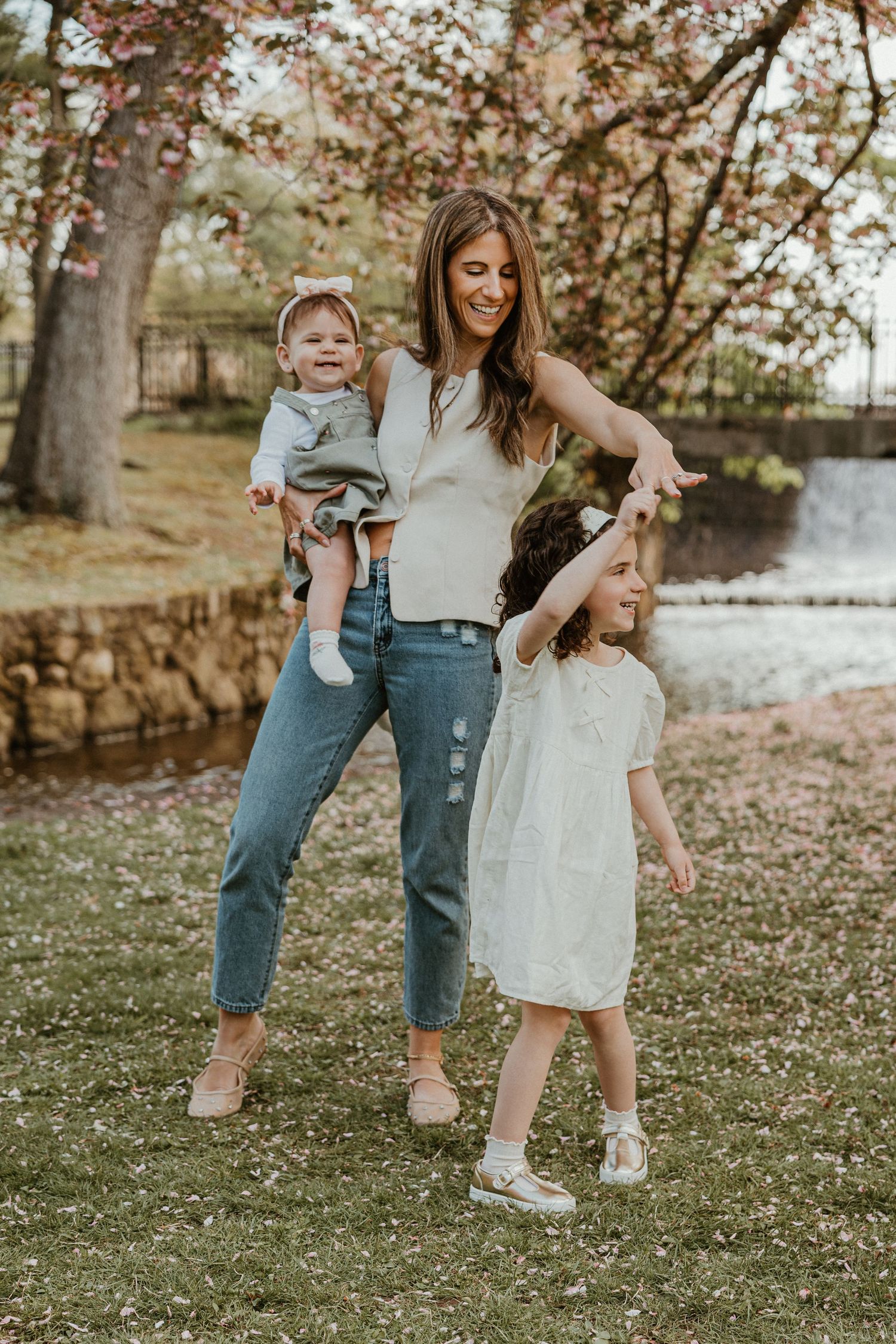 A smiling mother holds a baby while her young daughter dances beside her under blooming pink trees outdoors.