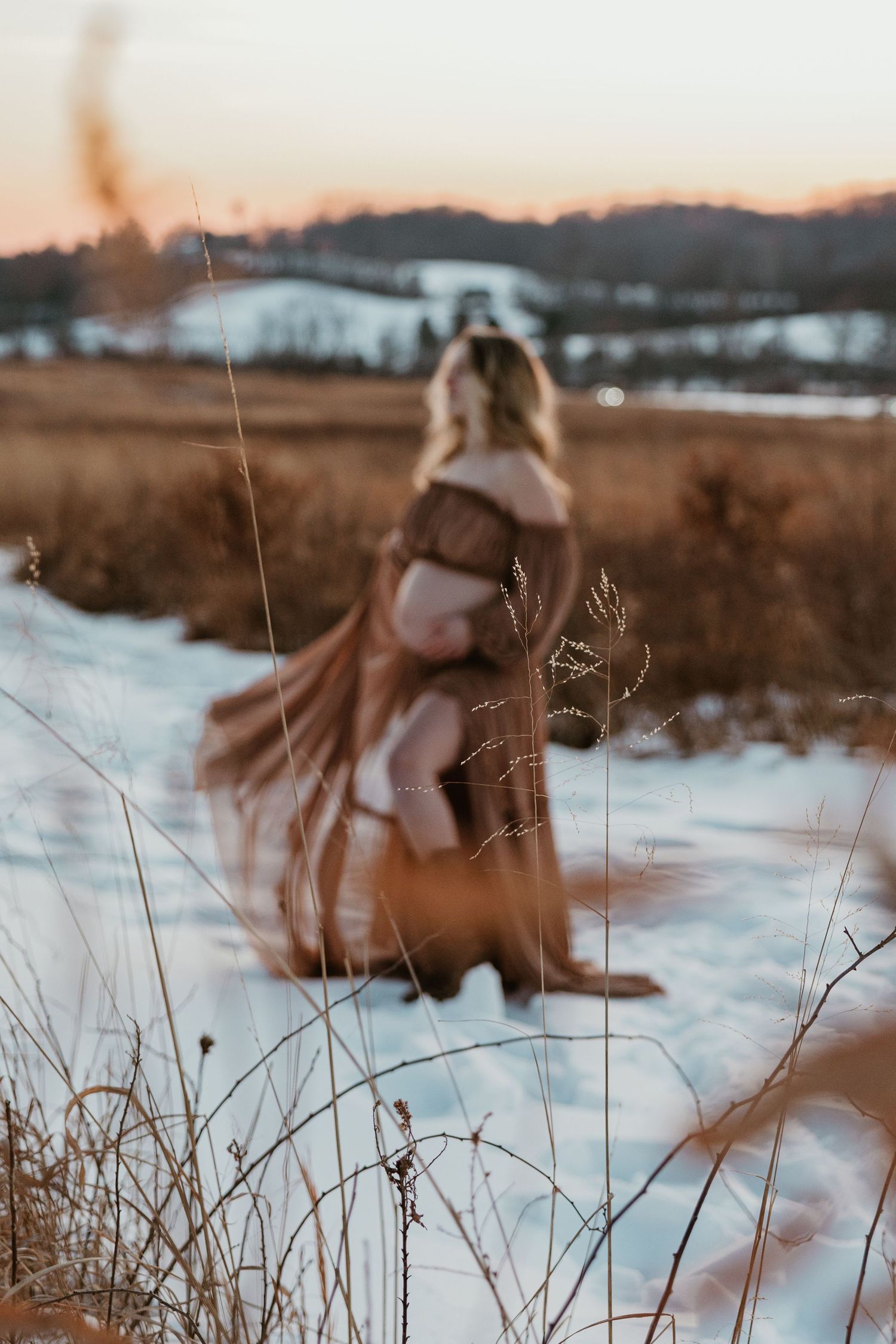 Artistic maternity silhouette in flowing brown dress against winter sunset landscape.