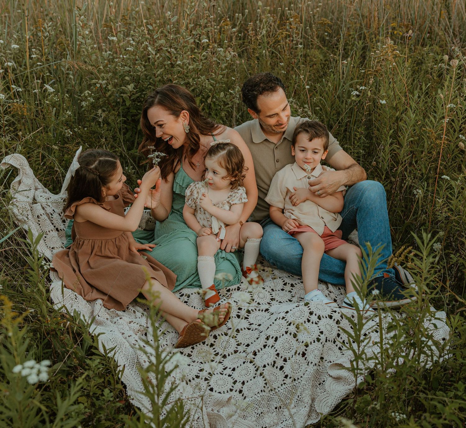 Family of five sitting on lace blanket in wildflower field, parents with three young children laughing together outdoors.