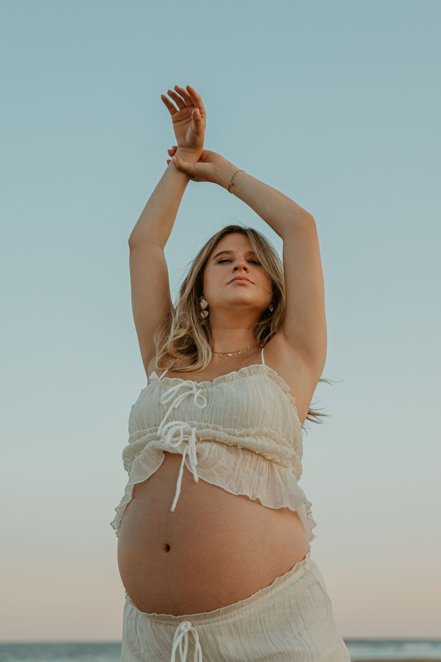 Pregnant woman in white crop top and pants poses with arms raised at beach during golden hour sunset.
