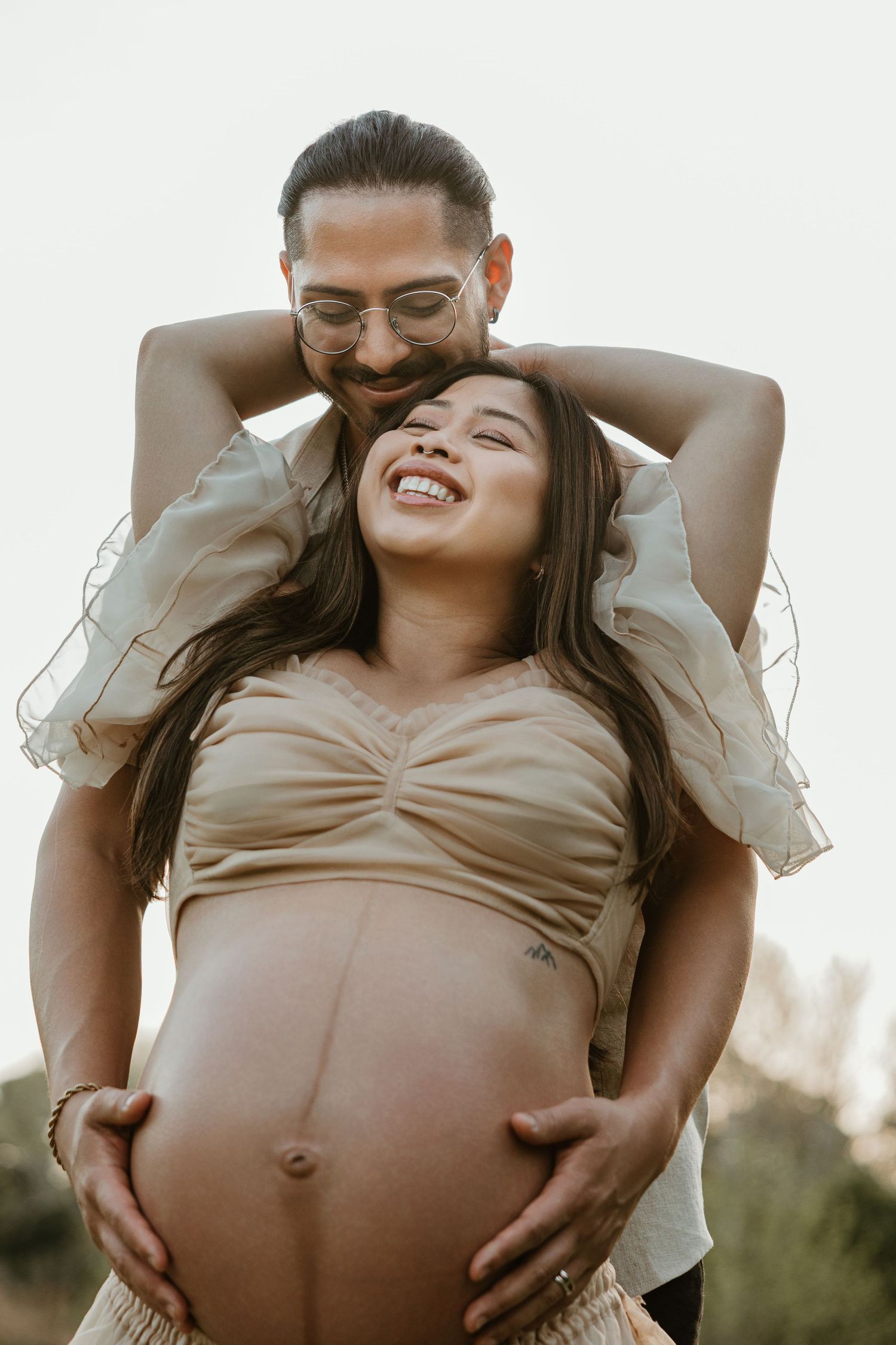 A smiling pregnant woman in a beige crop top is embraced from behind by a man outdoors in a warm maternity photo.