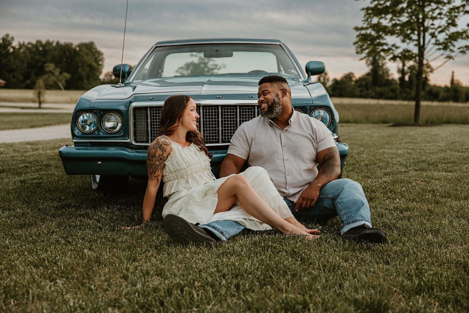 Couple sitting on grass leaning against a vintage teal car, smiling at each other during a golden hour outdoor photo session.