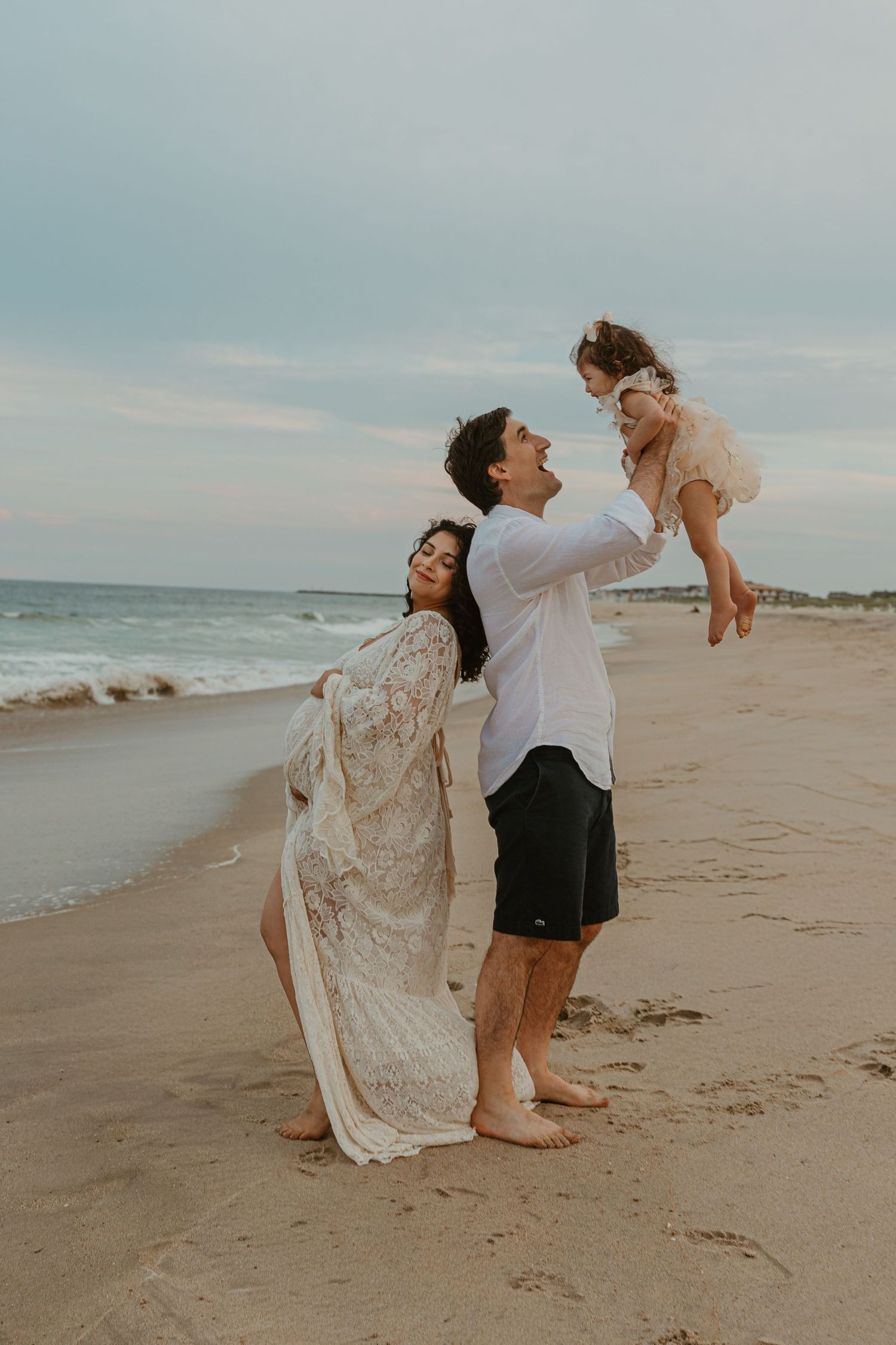 Family enjoying a beach day, man lifting toddler girl in air while woman in lace dress watches, ocean backdrop.