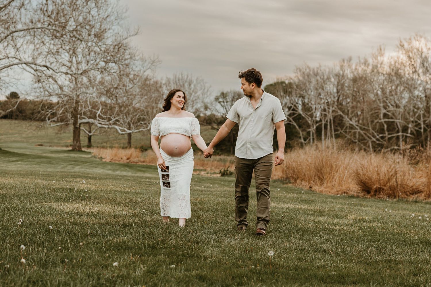 Pregnant couple holding hands and walking in a green park, woman holding ultrasound photo during outdoor maternity photoshoot.