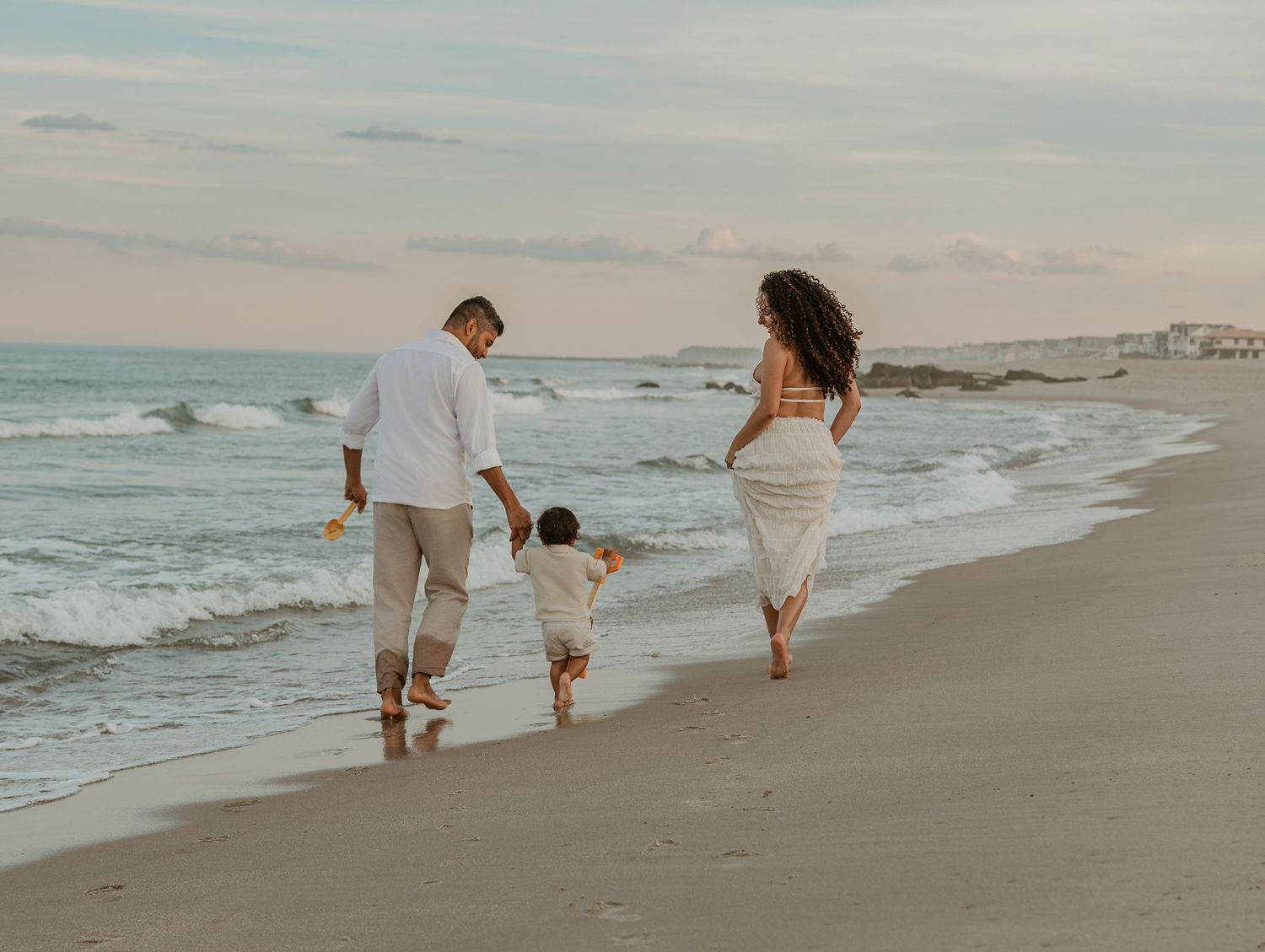Family of three walking hand-in-hand along a sandy beach at sunset, waves gently lapping the shore.
