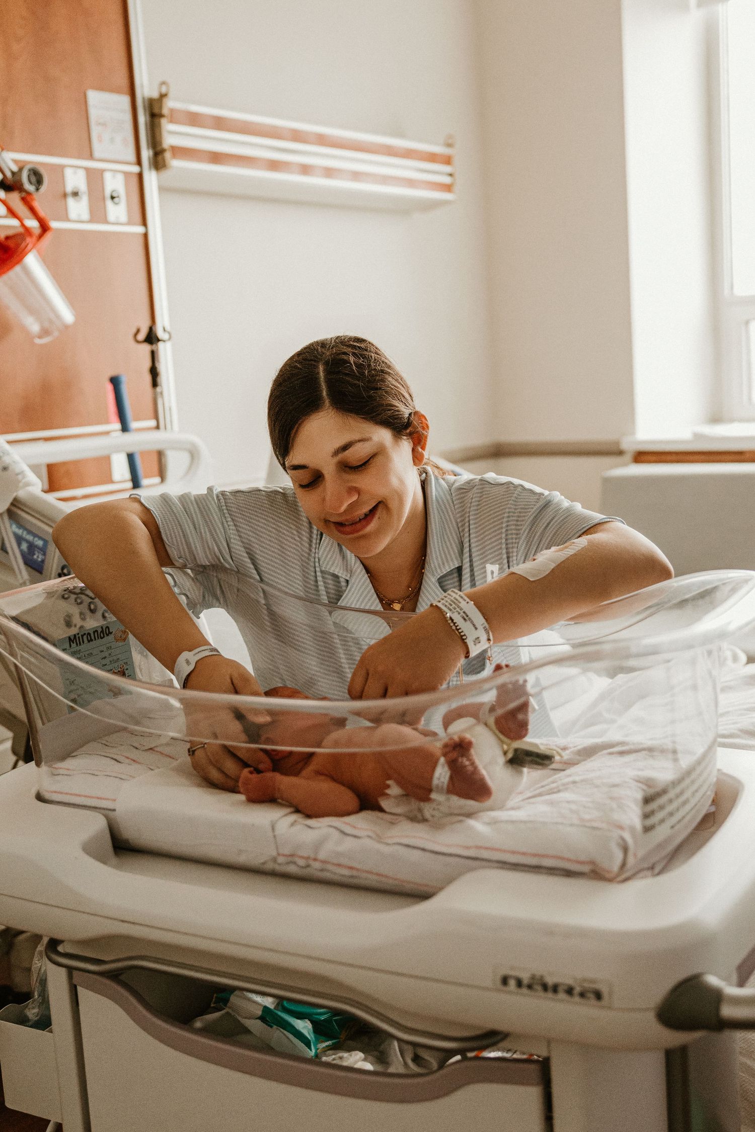 New mother smiling while reaching into hospital bassinet to hold her newborn baby.
