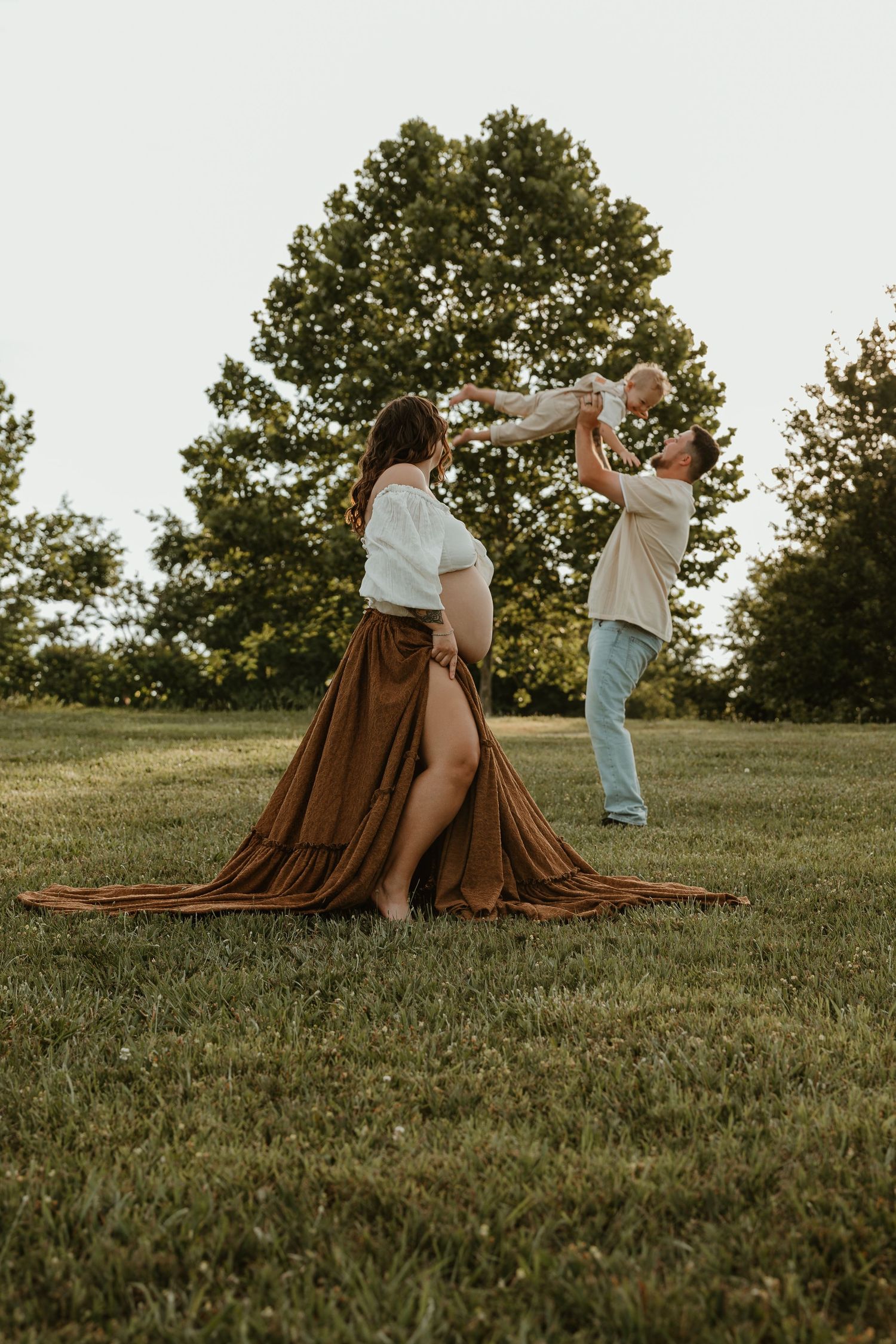Pregnant woman in brown skirt with partner lifting baby outdoors on green grass with trees in background.