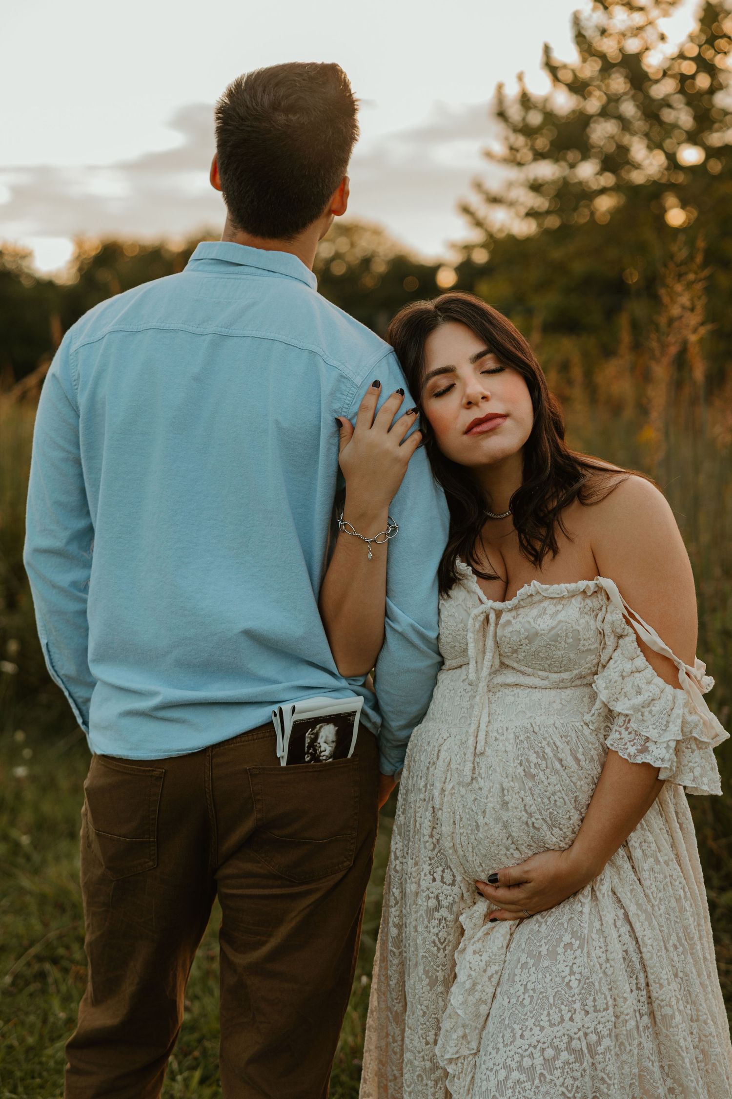 Pregnant woman in white lace dress leans on partner holding ultrasound photo during golden hour maternity shoot.