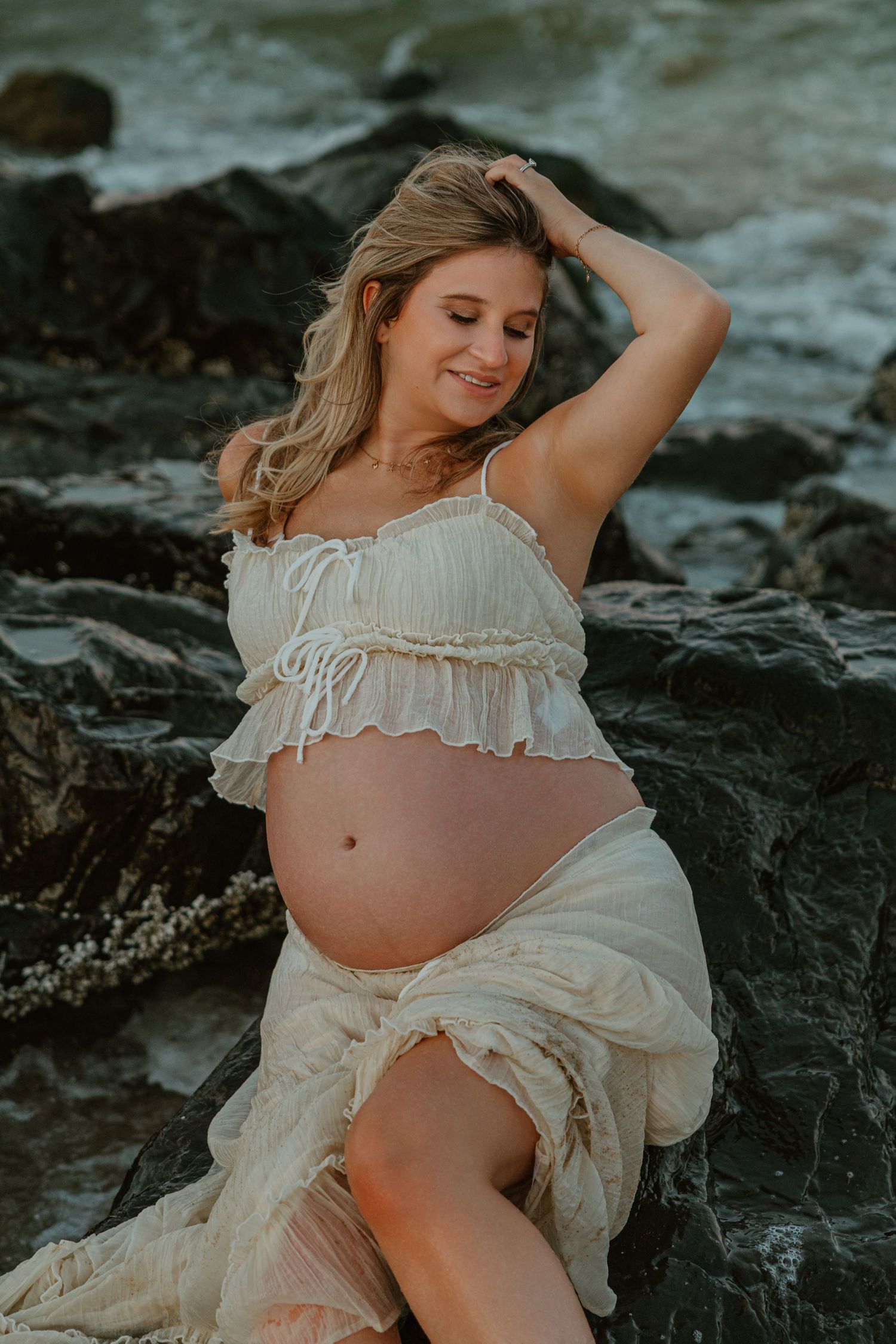 Pregnant woman in white crop top and sheer skirt sits on rocks by ocean, smiling and touching her hair.
