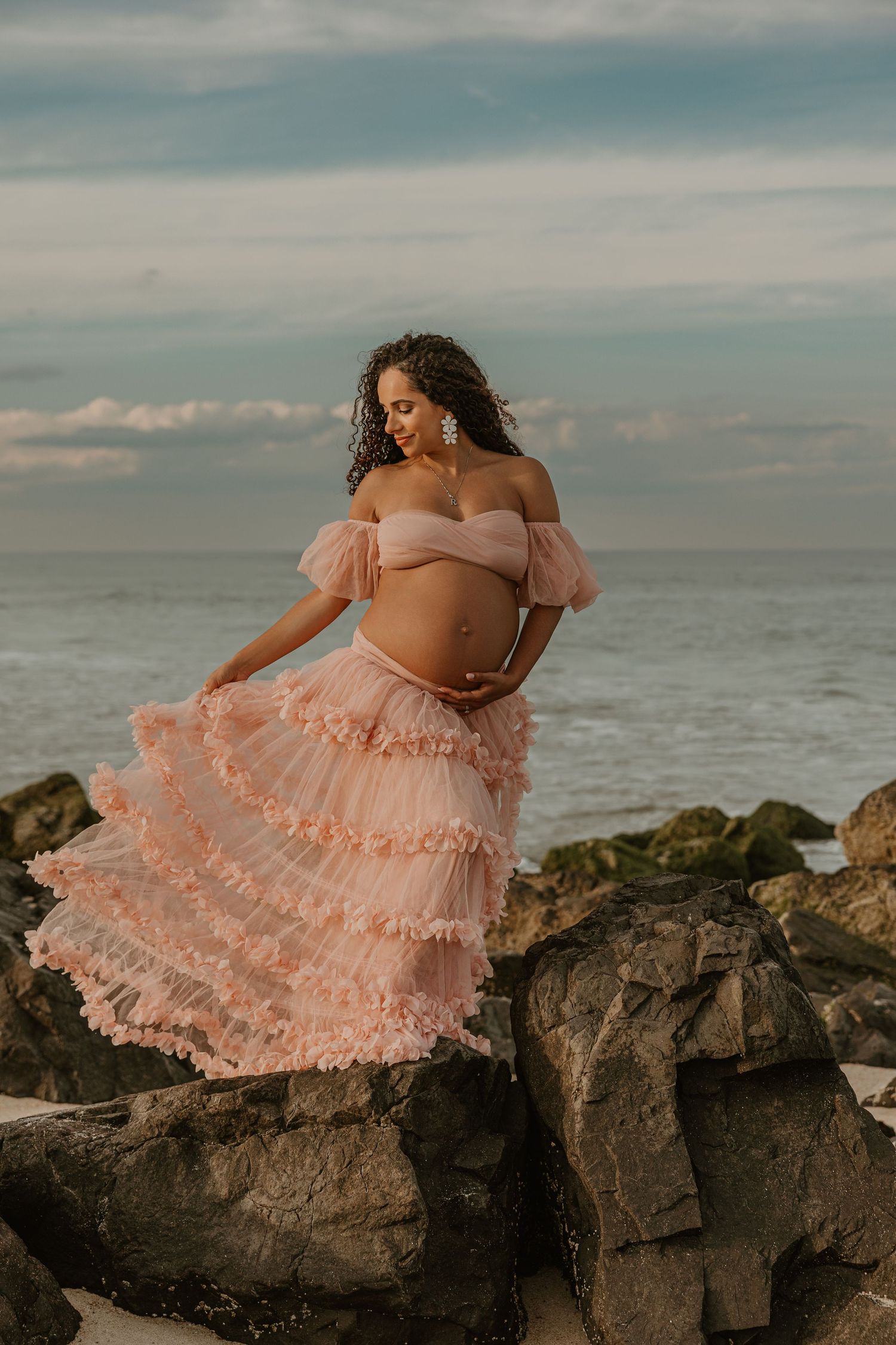 Pregnant woman in pink ruffled off-shoulder gown poses on rocky beach with ocean backdrop.