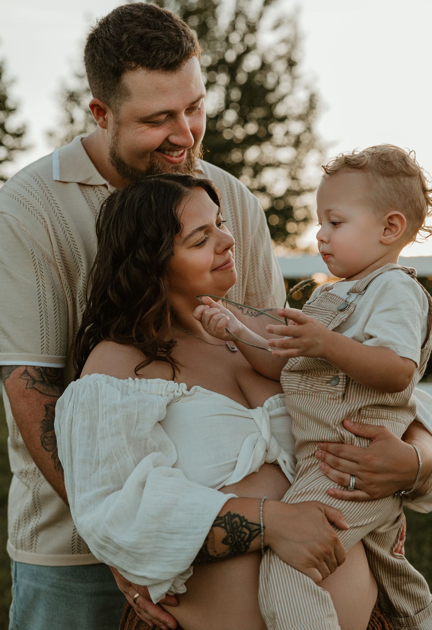 A smiling family with tattoos enjoys a golden hour outdoor moment, as a toddler boy reaches toward his pregnant mother.