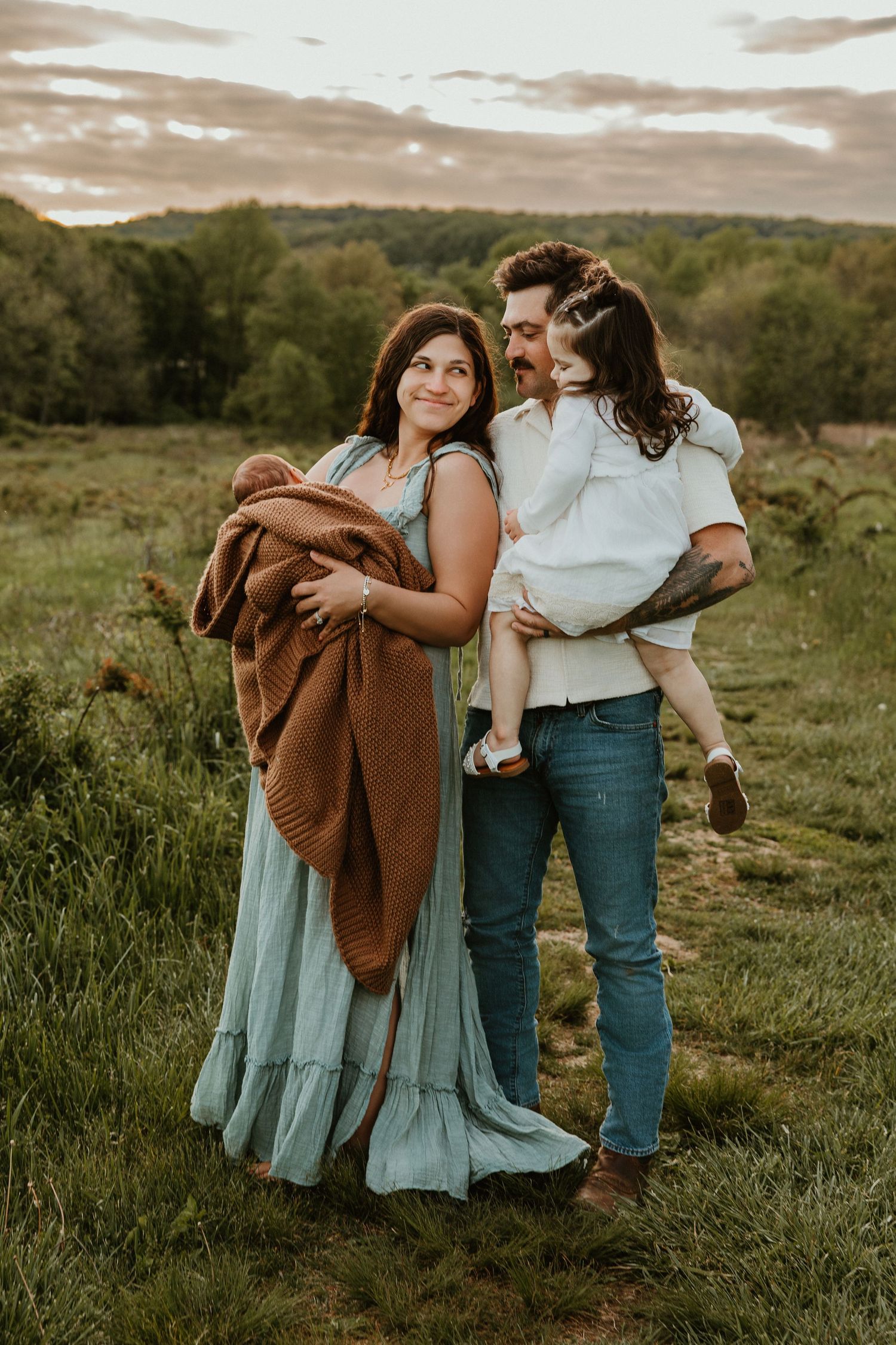 Family portrait in a green meadow at sunset, woman in teal dress holding newborn, man carrying toddler.