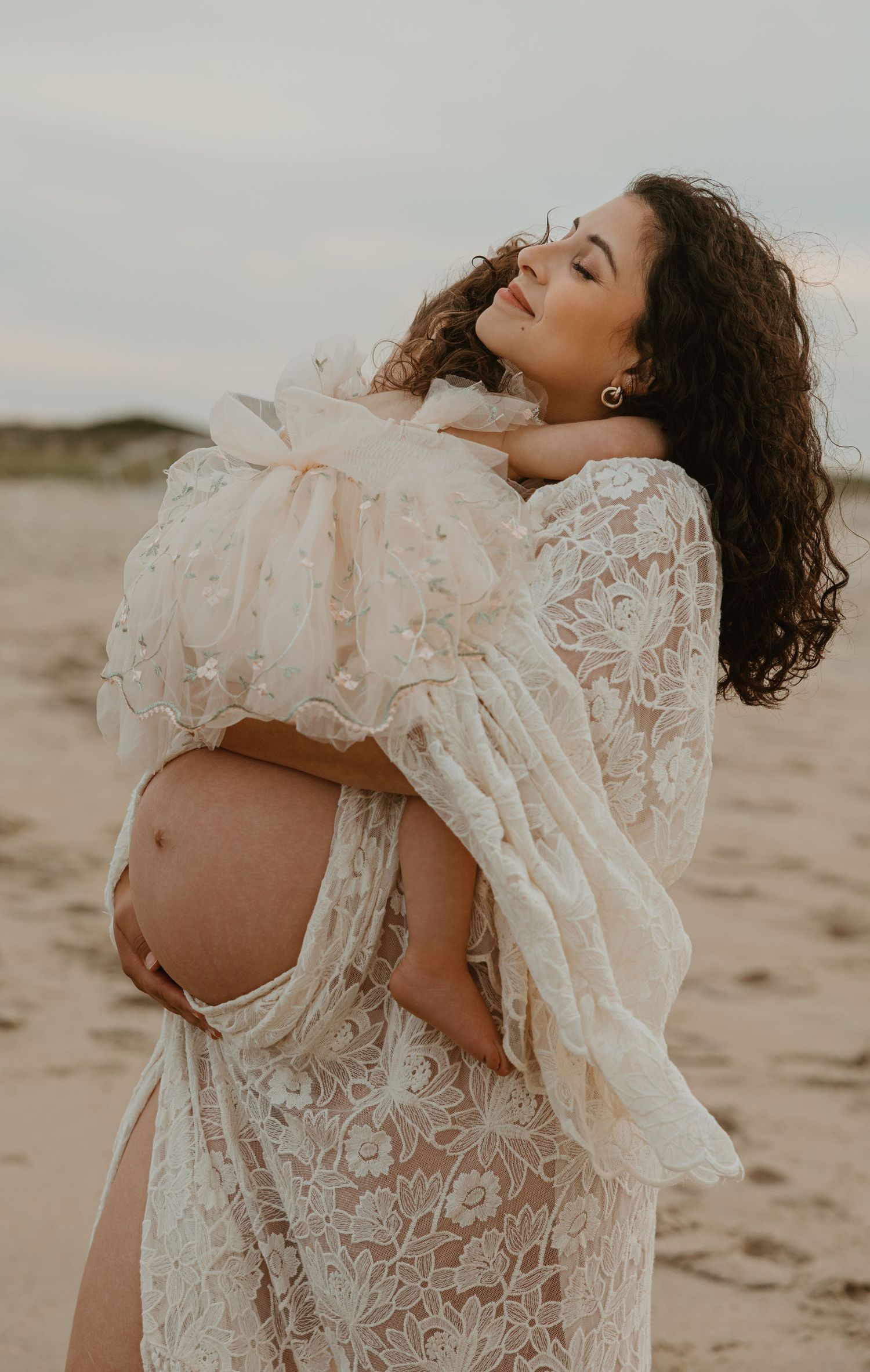 Pregnant woman in white lace dress cradling baby bump on sandy beach at dusk.