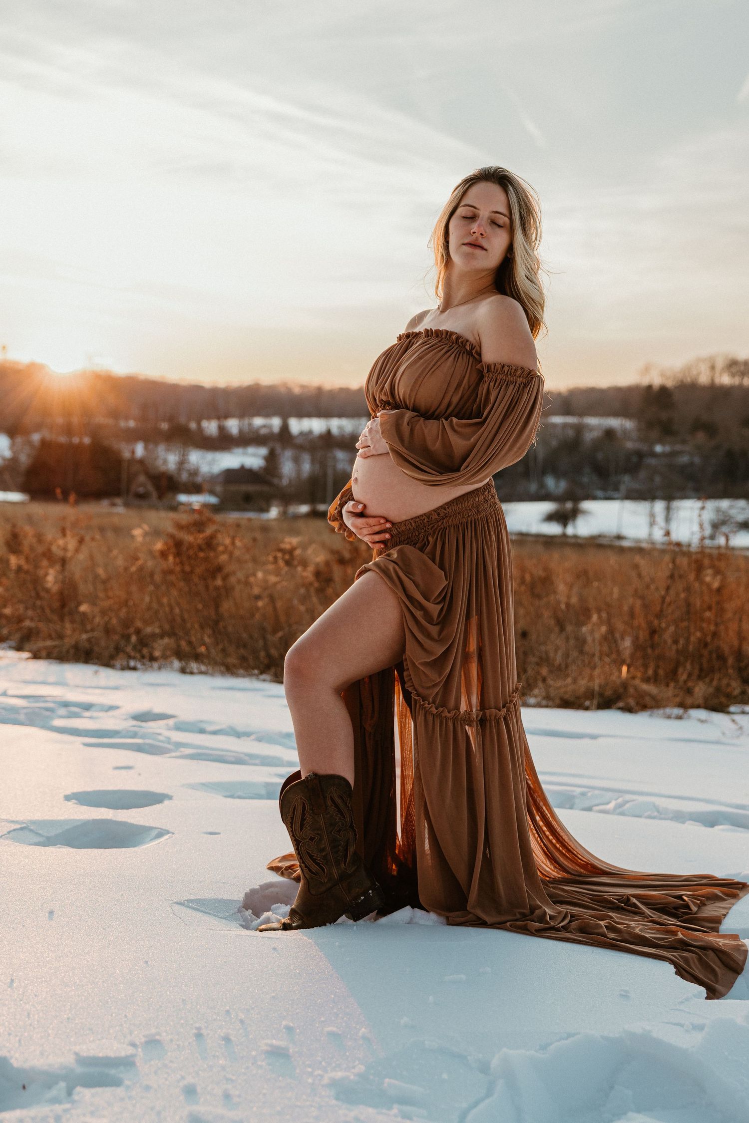 Winter maternity session captures golden hour light on snow with flowing brown fabric.