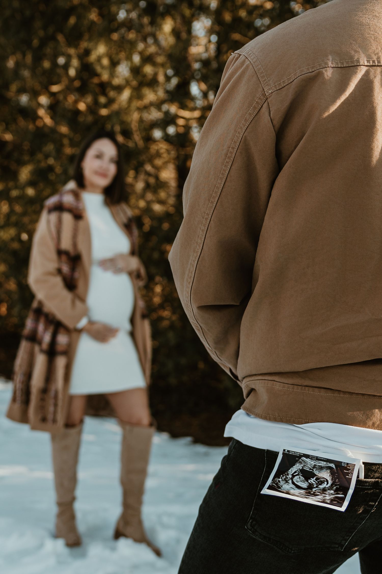 A pregnancy announcement photoshoot in winter shows a couple holding ultrasound photos in the snow.