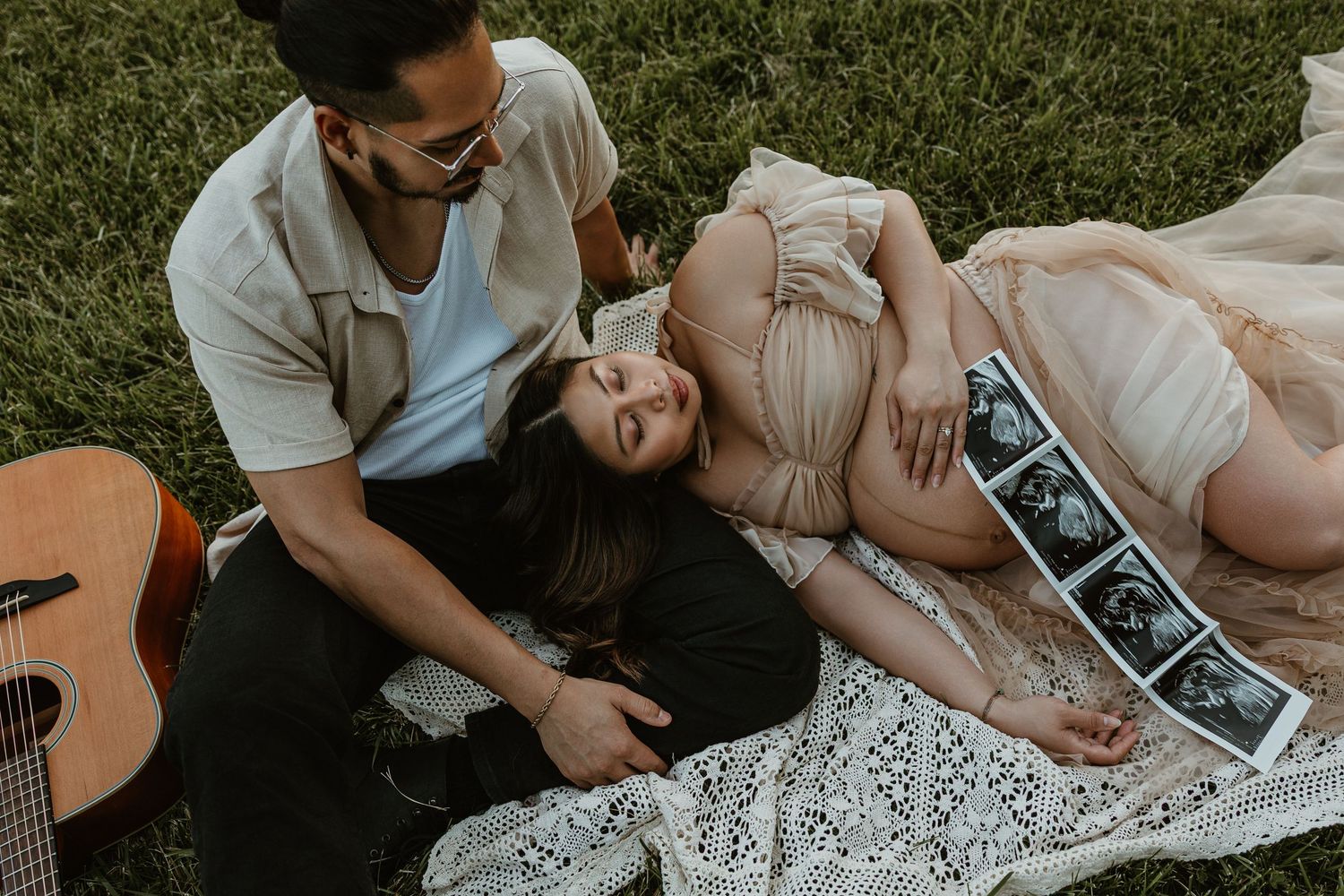 Couple sharing a tender moment on a blanket in the grass with ultrasound photos and a guitar nearby.