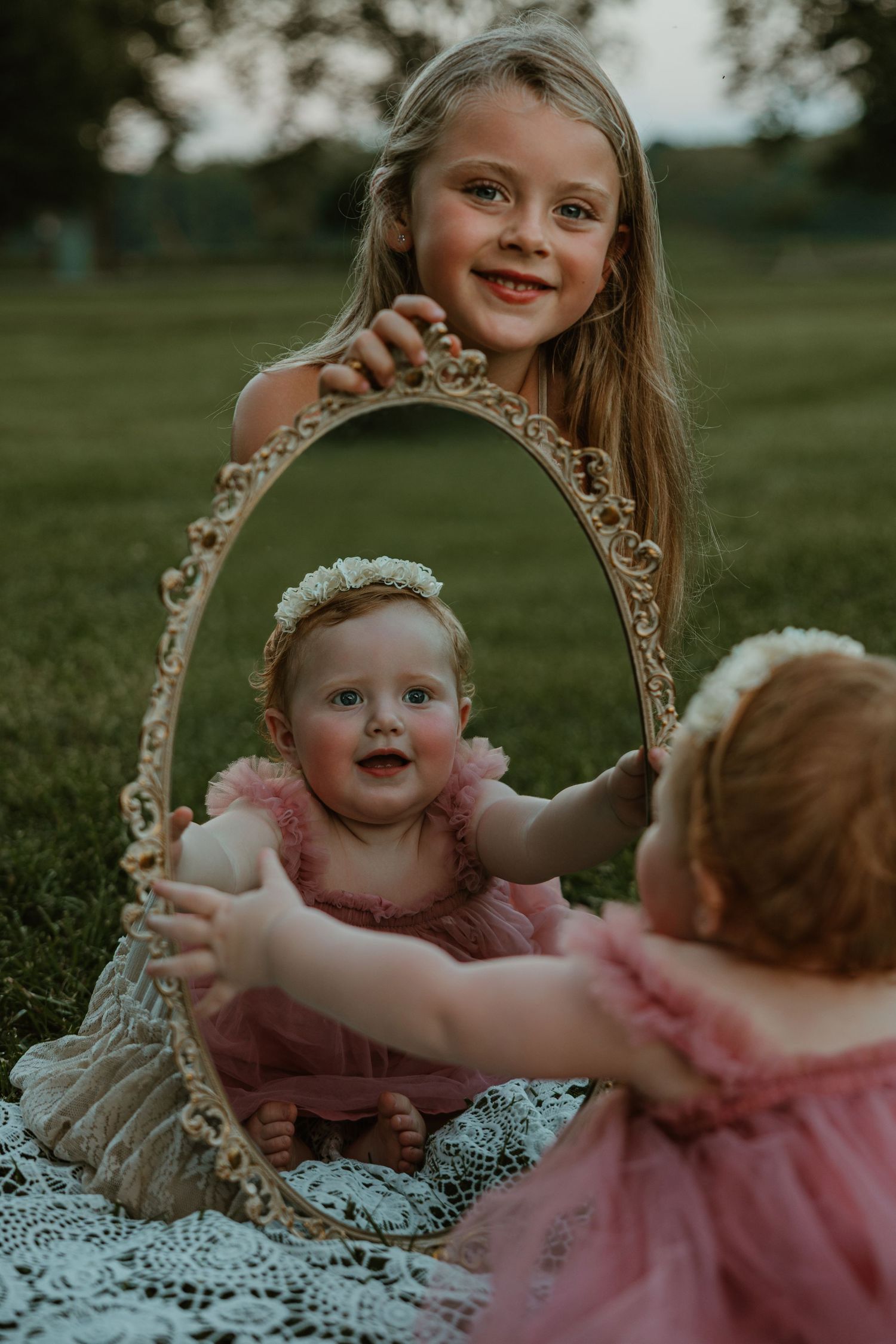 A smiling girl holds an ornate mirror reflecting a baby in a pink dress outdoors on a lace blanket.