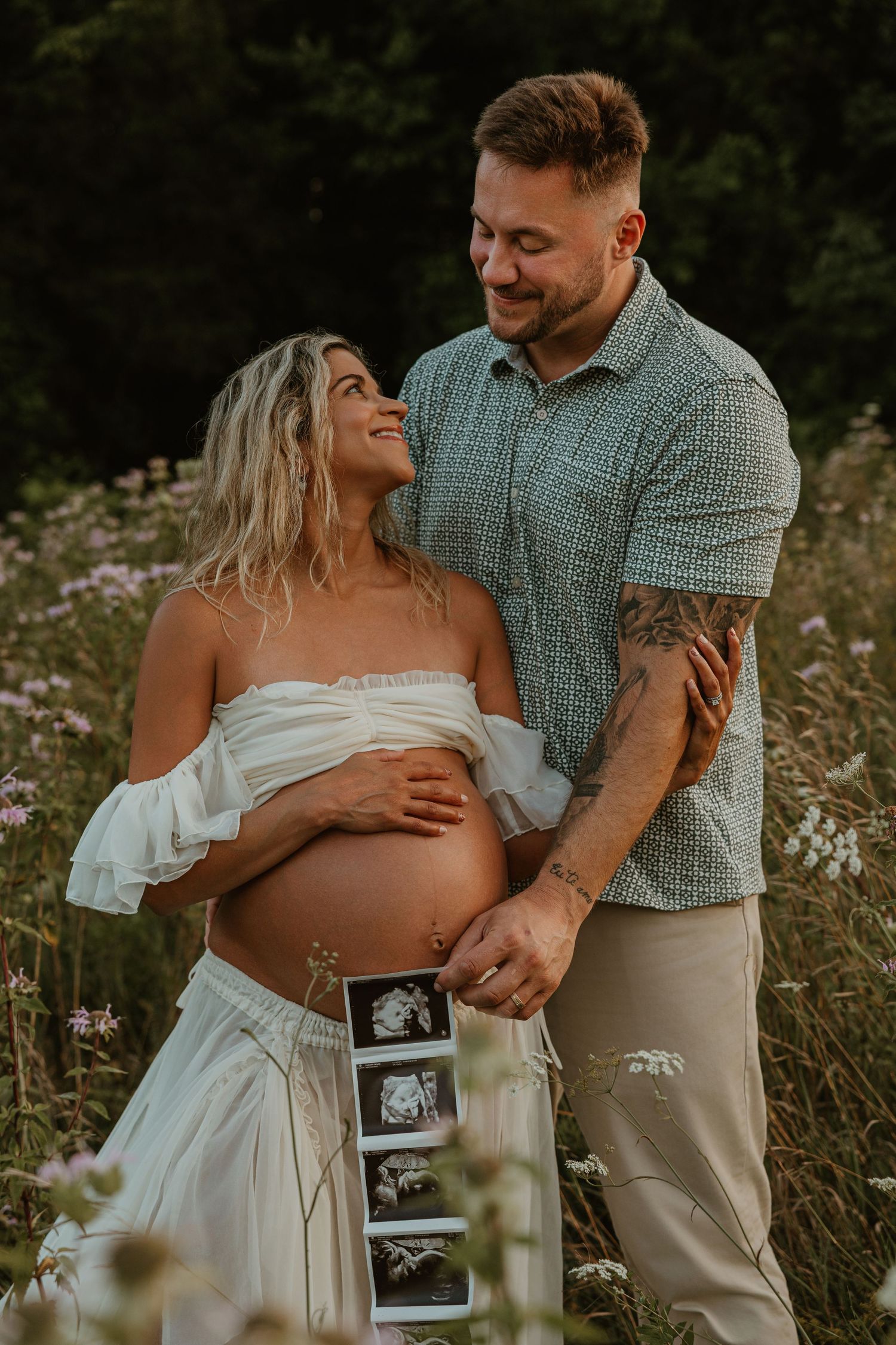 Couple in wildflower field holding ultrasound photos during maternity shoot.