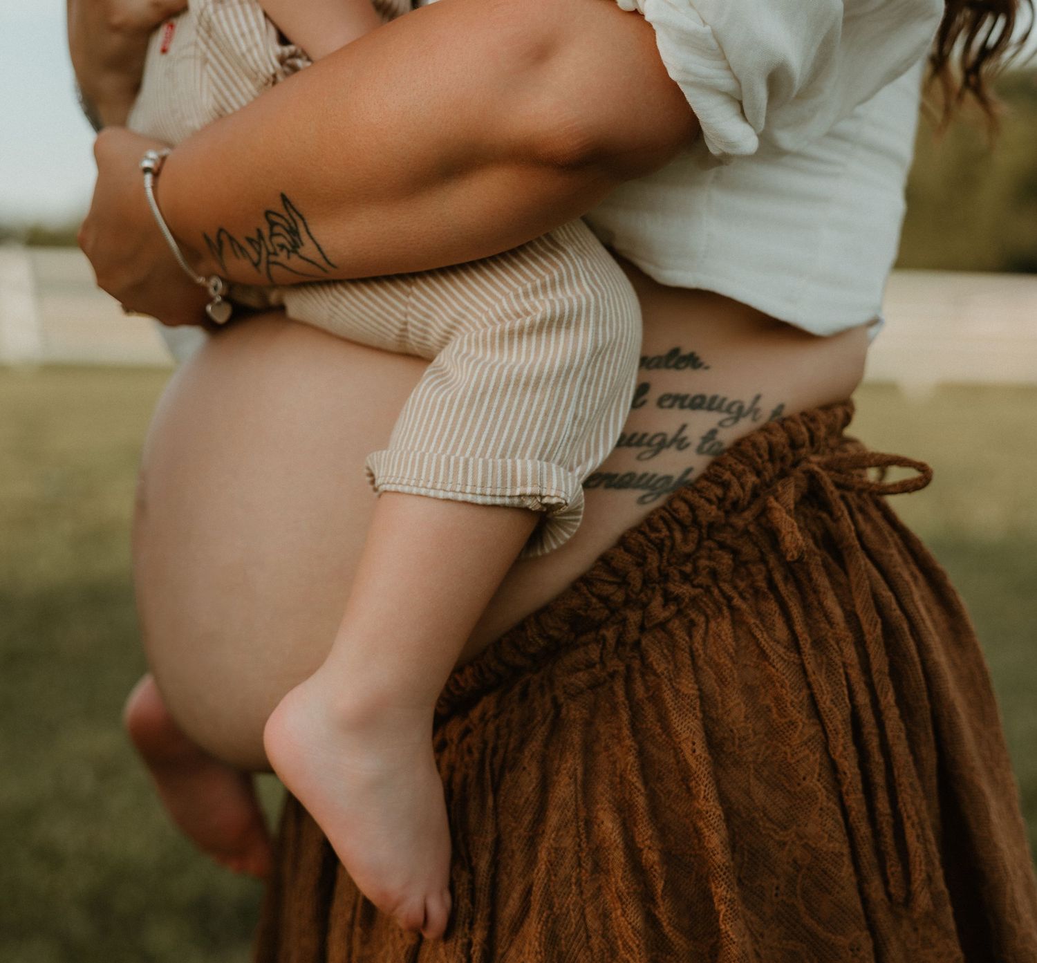 Mother with tattoos holding a baby dressed in striped shorts outdoors, wearing a brown skirt.