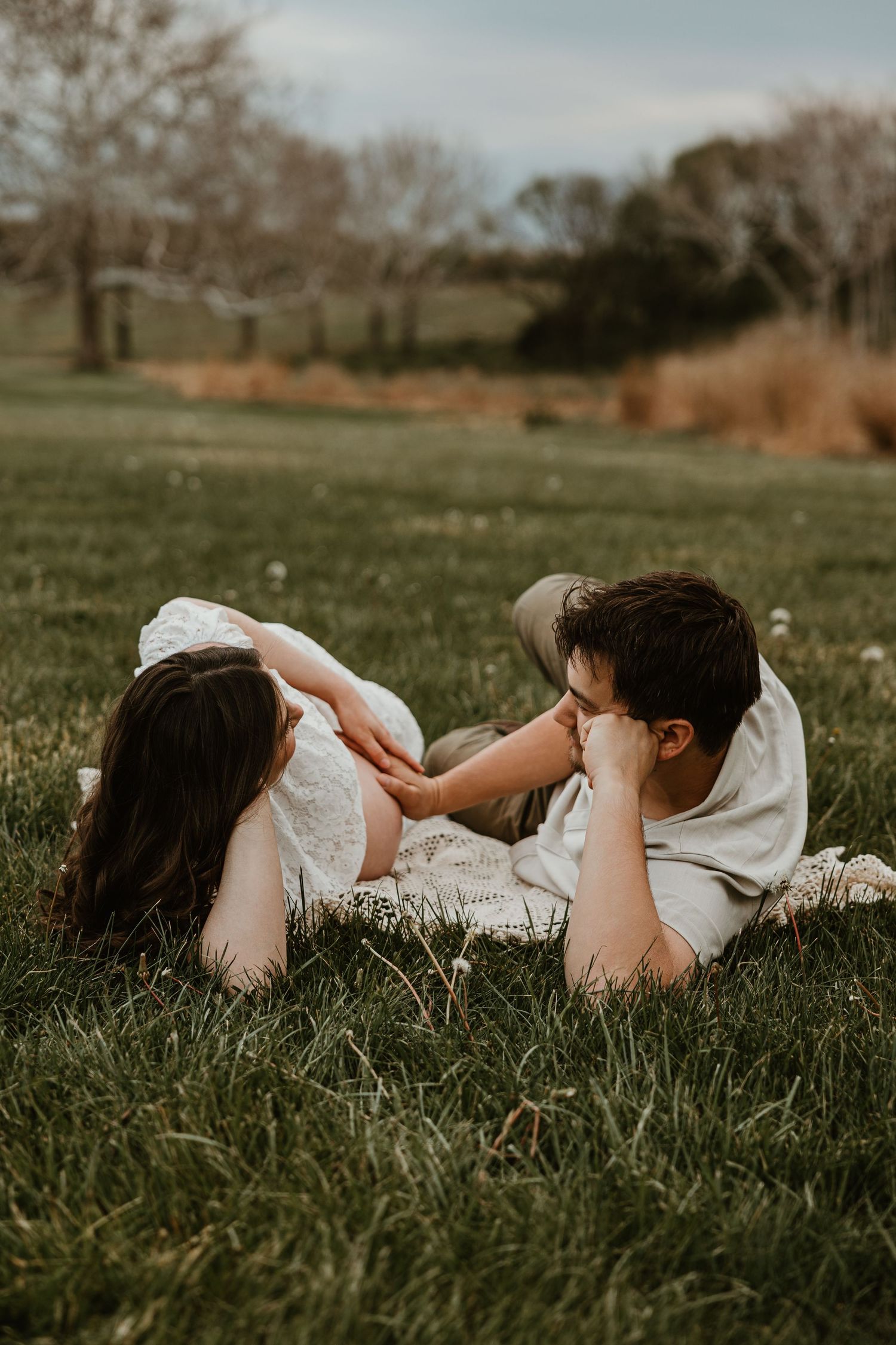 Couple lying on green grass in a field, sharing an intimate moment during a romantic outdoor maternity session.