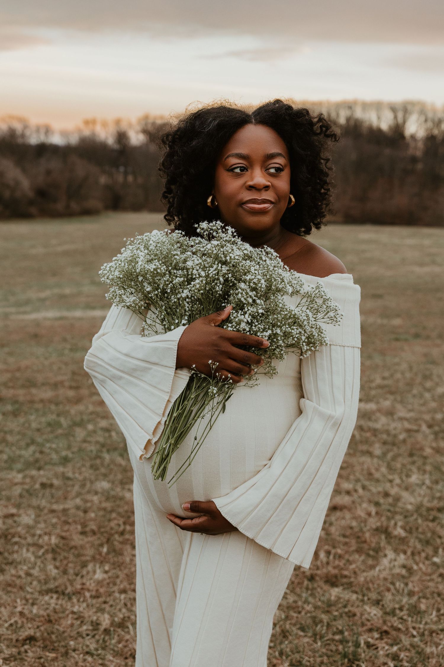 Pregnant Black woman in white off-shoulder dress holding baby's breath flowers in a field at sunset.