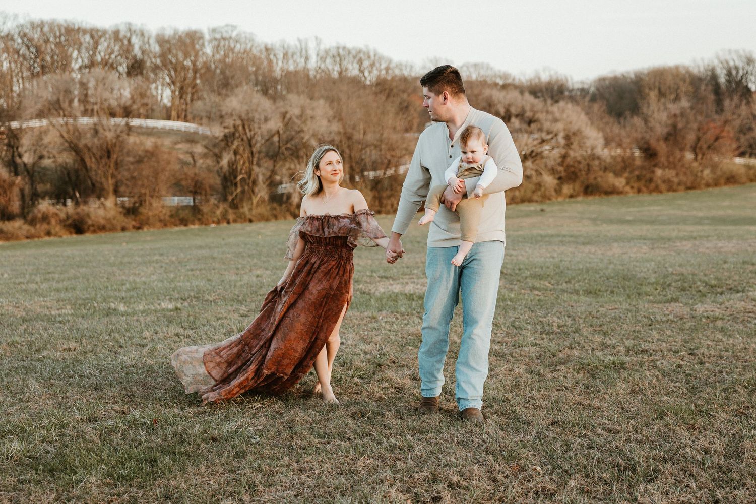 A young family poses outdoors in a field, mom in a flowing floral dress, dad holding a baby, during golden hour.
