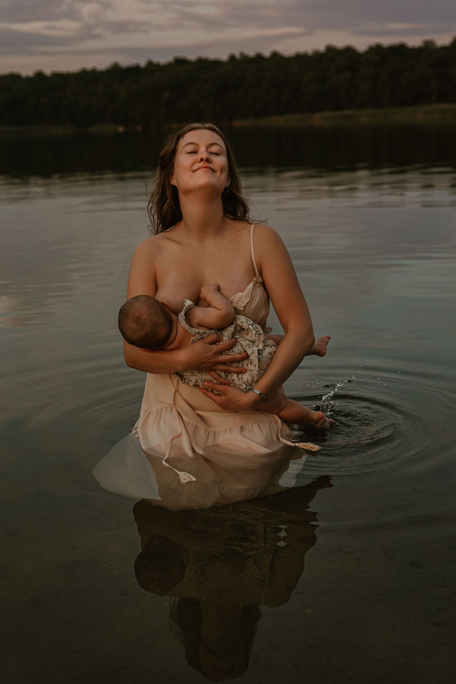 Mother breastfeeding infant while standing in calm lake at dusk, wearing a light dress with forest in background.