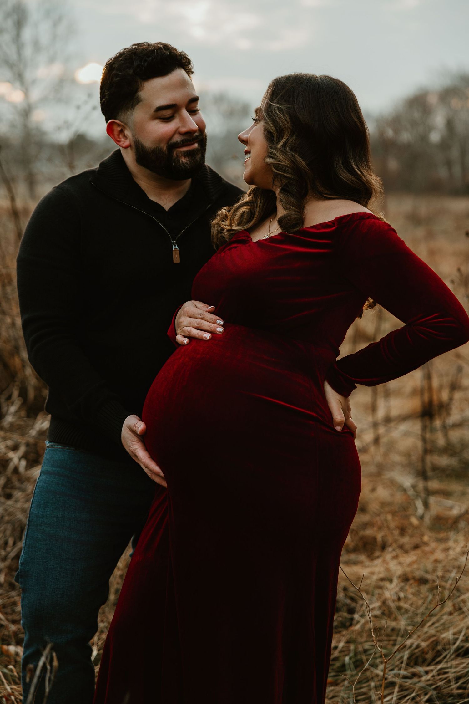 Couple in black and burgundy outfits embrace in golden autumn field.