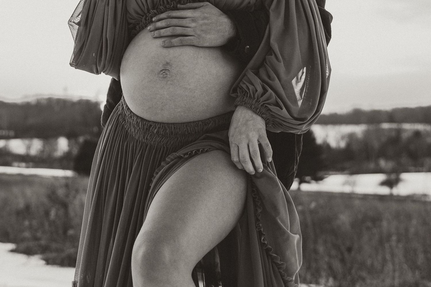 Black and white detail shot of maternity pose in flowing dress against winter landscape.