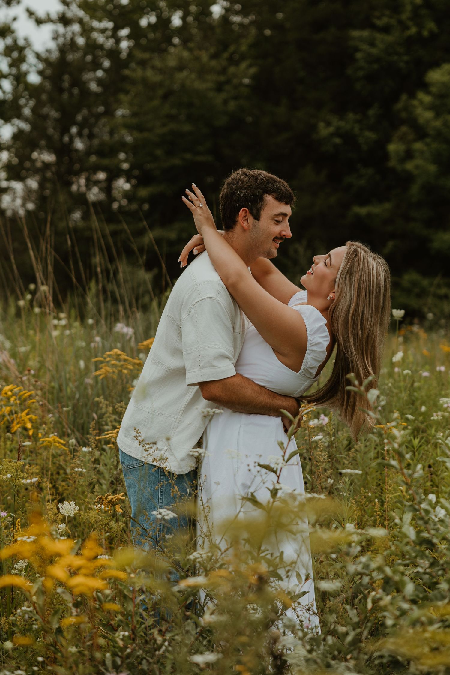 A couple embraces and laughs together in a wildflower meadow, wearing white and denim outfits with trees in the background.