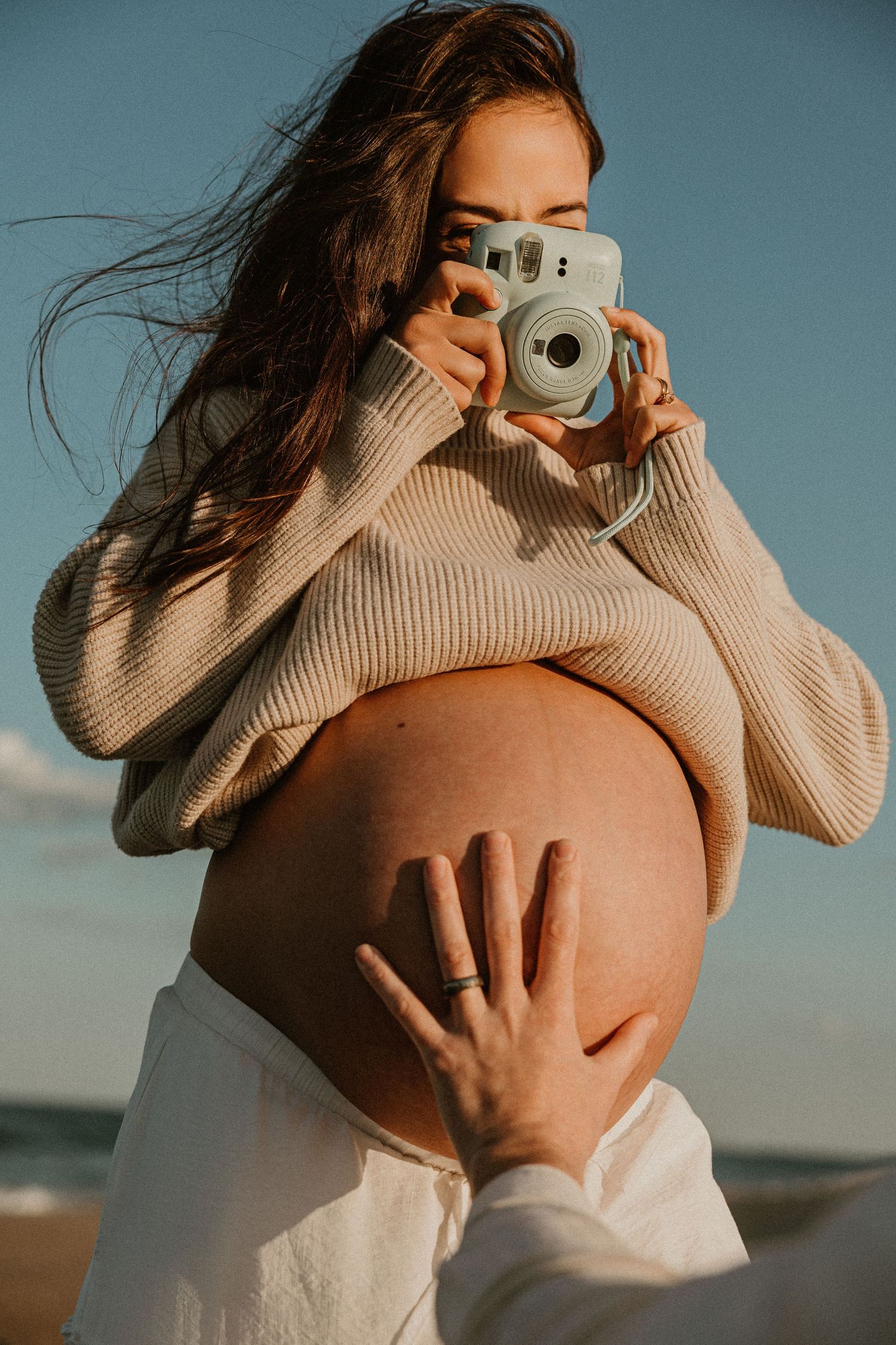 Pregnant woman in beige sweater holding Instax camera at beach, cradling bare baby bump outdoors.