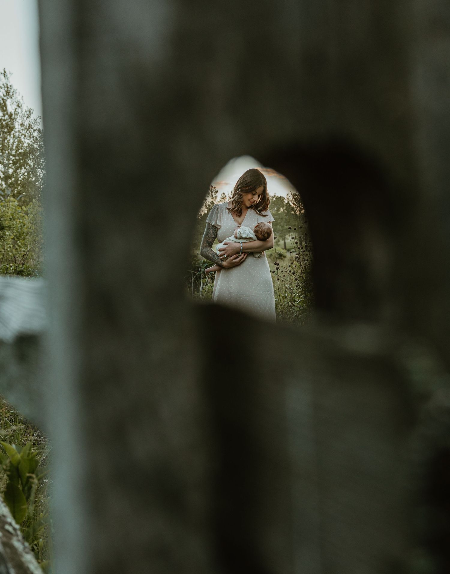 A bride in a white lace gown holds her newborn baby, framed by a stone archway surrounded by lush greenery.