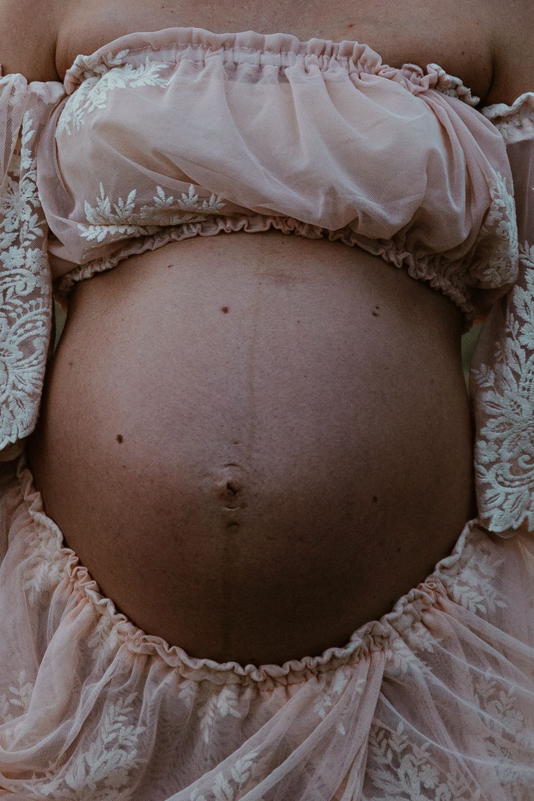 Close-up detail of pregnant belly framed by delicate pink lace dress with ruffled edges.