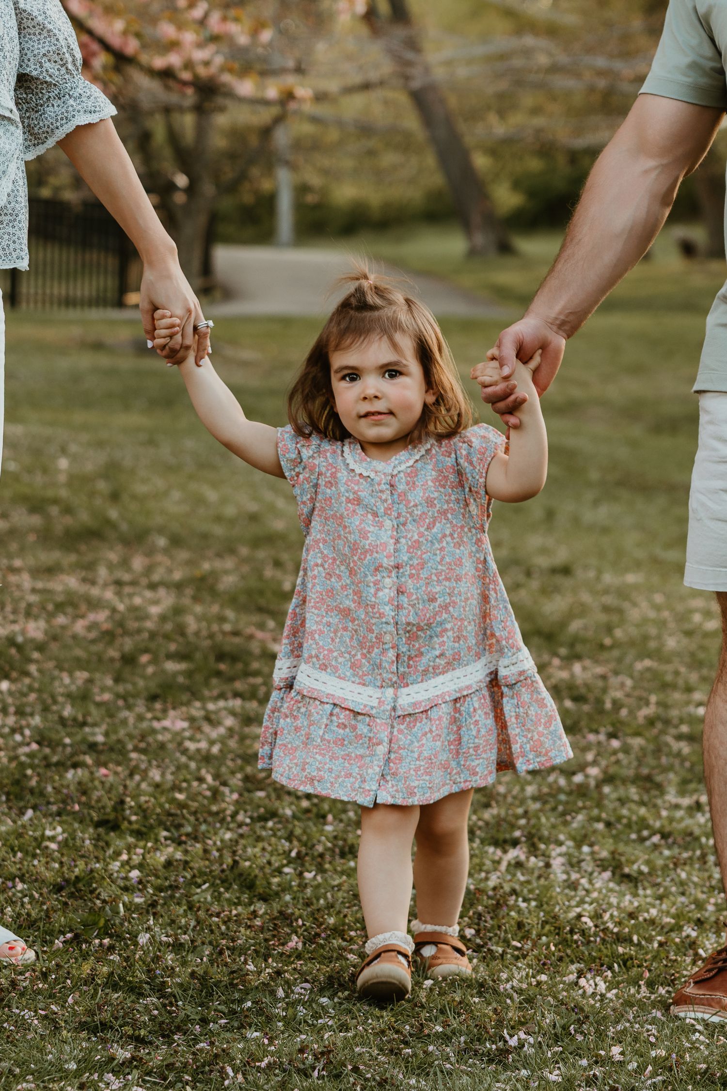Toddler girl in floral dress holds parents hands while walking in a blooming spring park.